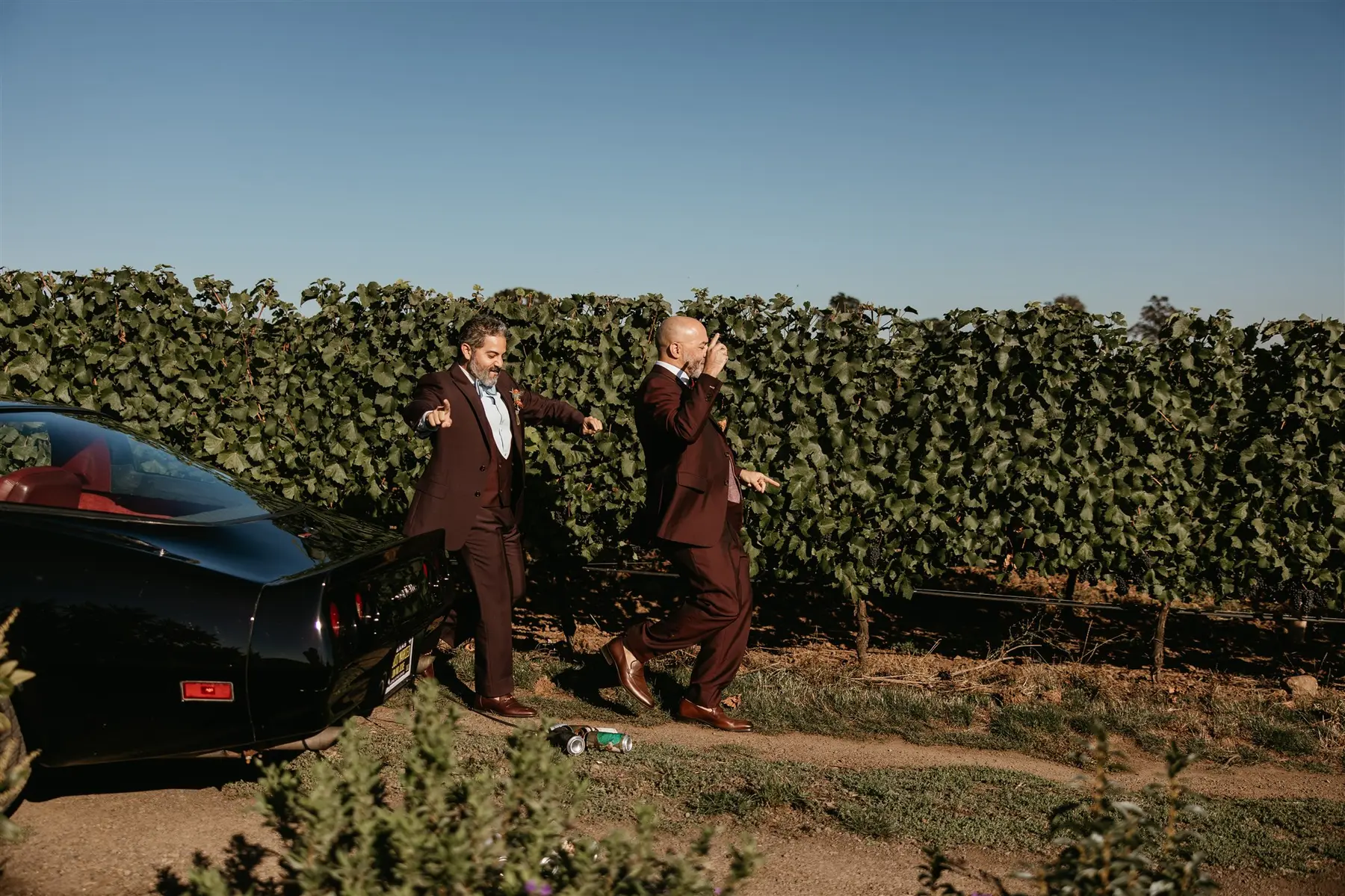 Josue Diaz and Jonathan Snowden in burgundy suits laugh and point at each other beside a black sports car at Stoller Family Estate.