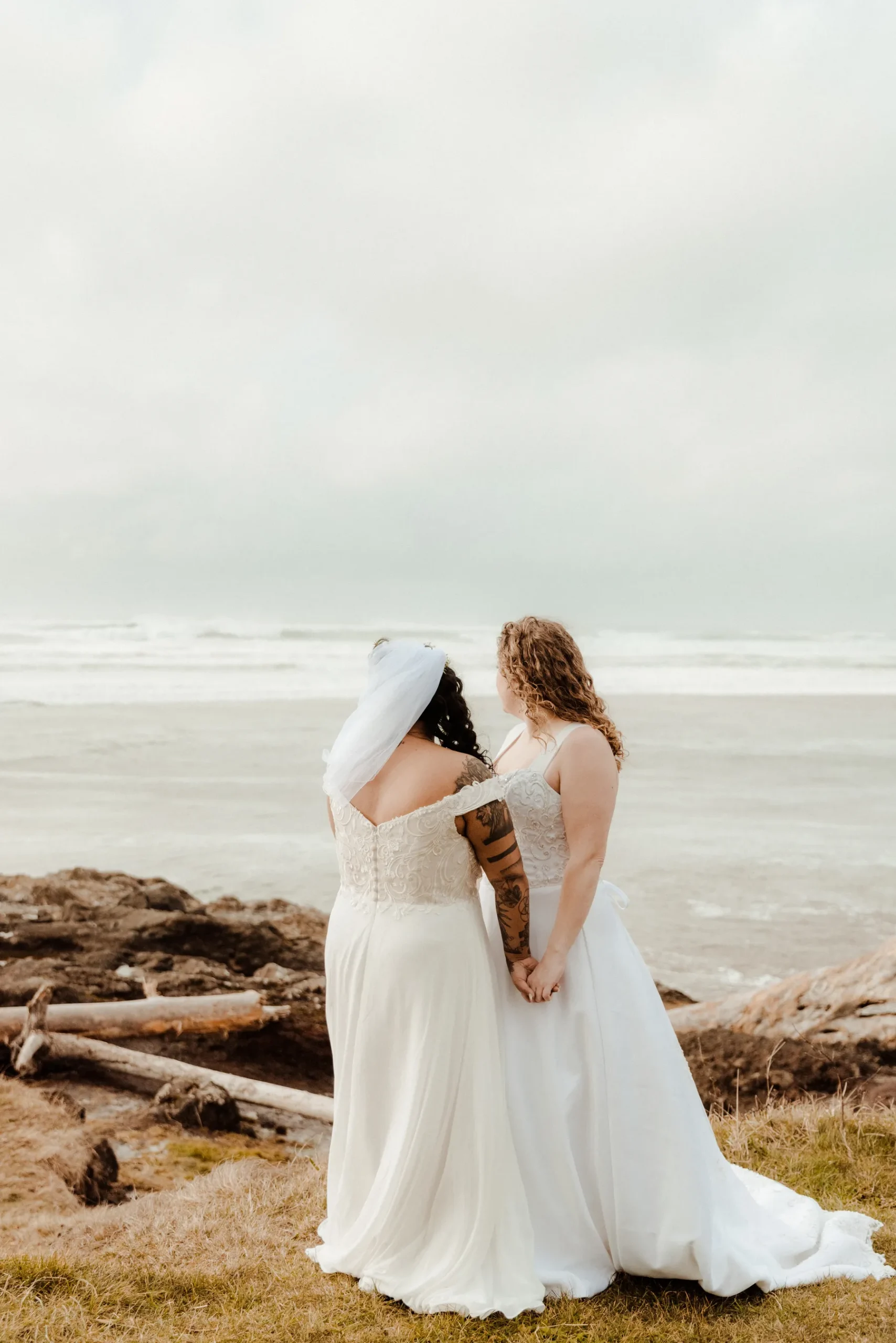 Brides Rachel and Jamie Snow hold hands facing the Pacific Ocean in white gowns at Yachats Inn, Oregon.