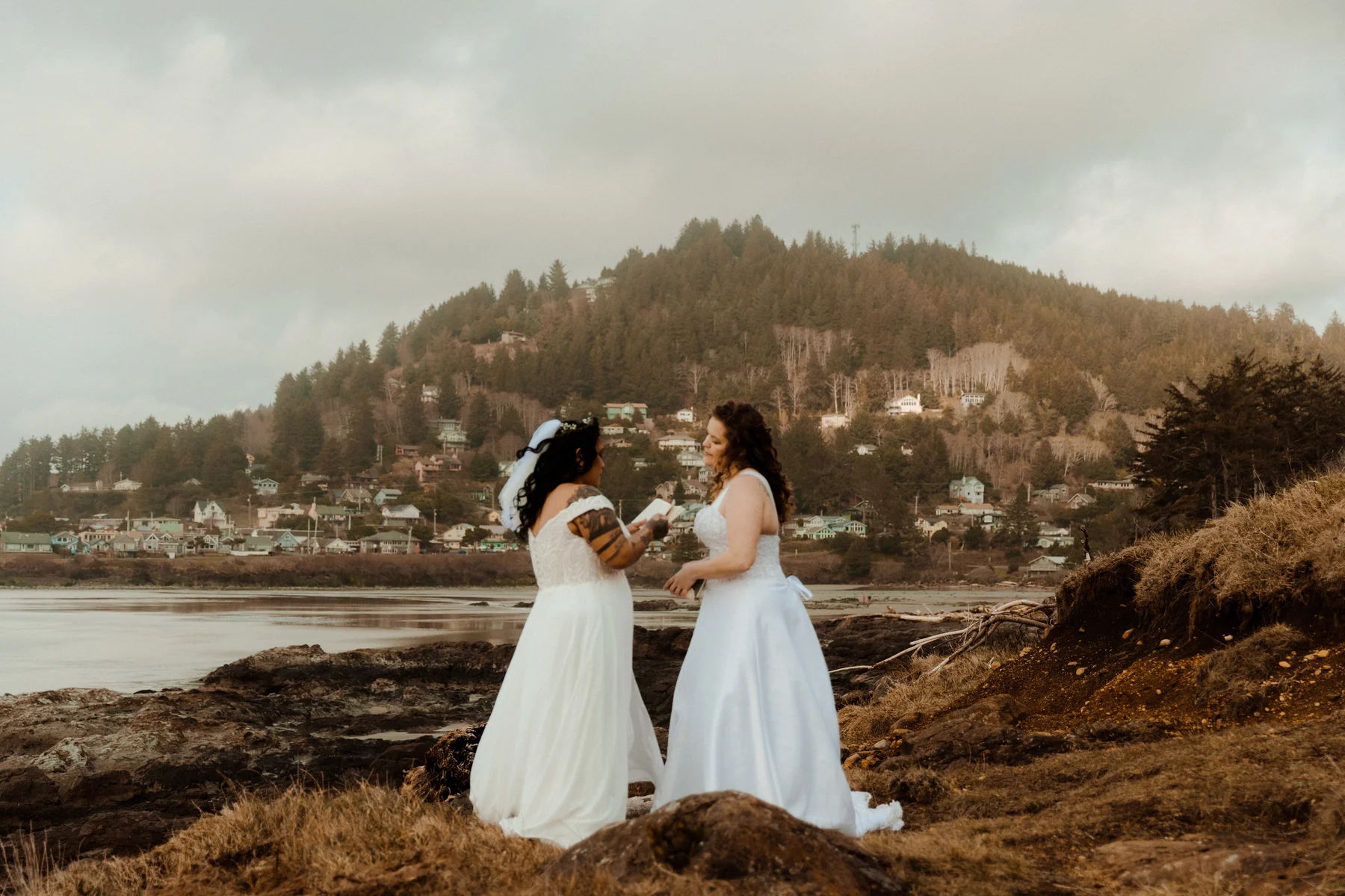 Brides Rachel and Jamie Snow exchange vows on a rocky Oregon coastline with a forested hill and coastal town behind them.
