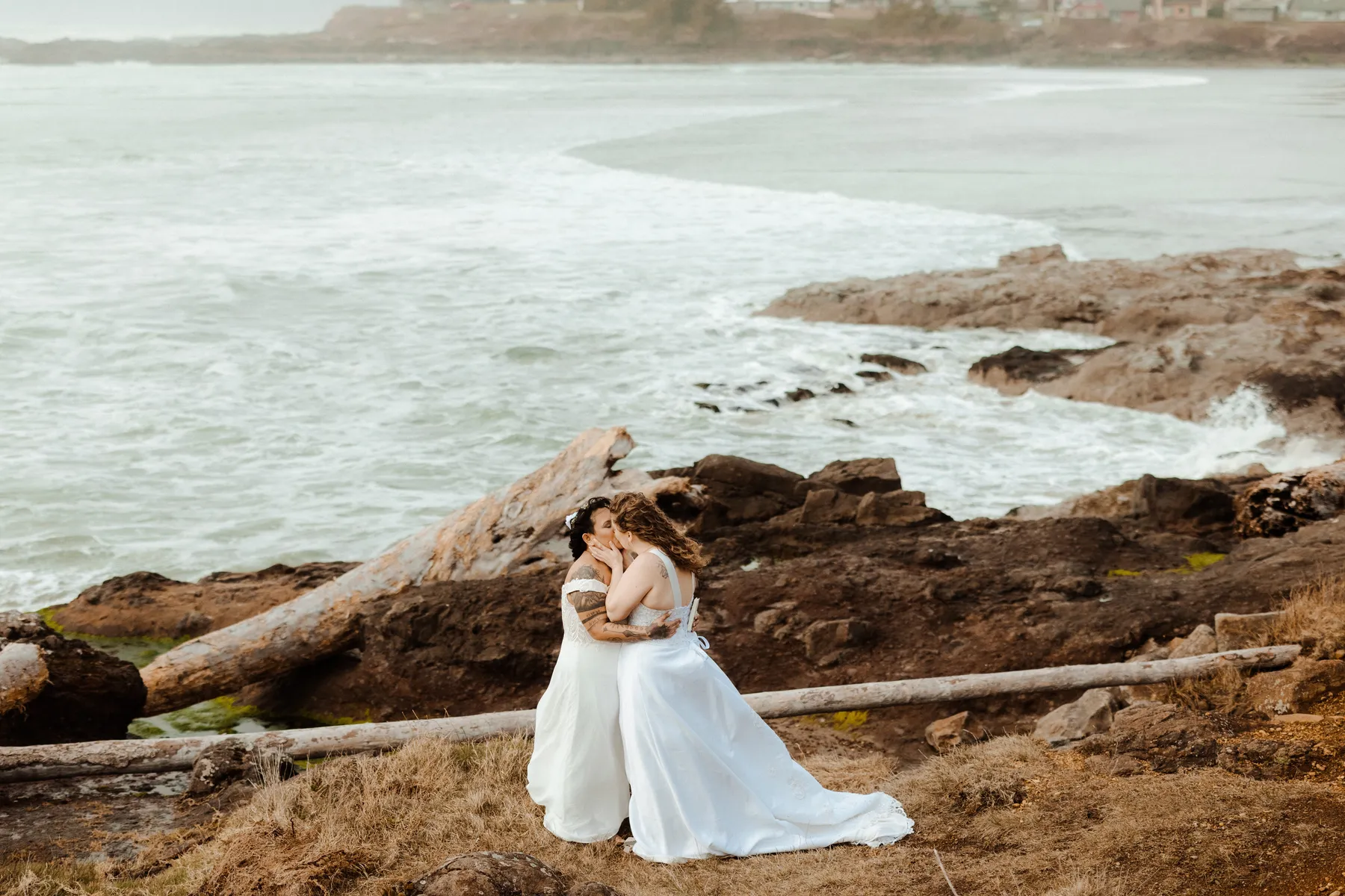 Brides Rachel and Jamie Snow share a kiss in white gowns on the rocky Oregon coast at Yachats Inn.