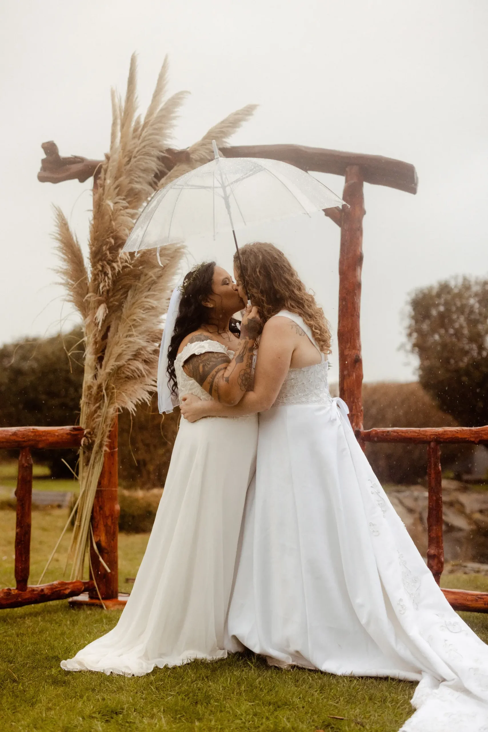 Brides Rachel and Jamie Snow share a first kiss under a clear umbrella at Yachats Inn in Oregon on a rainy wedding day.