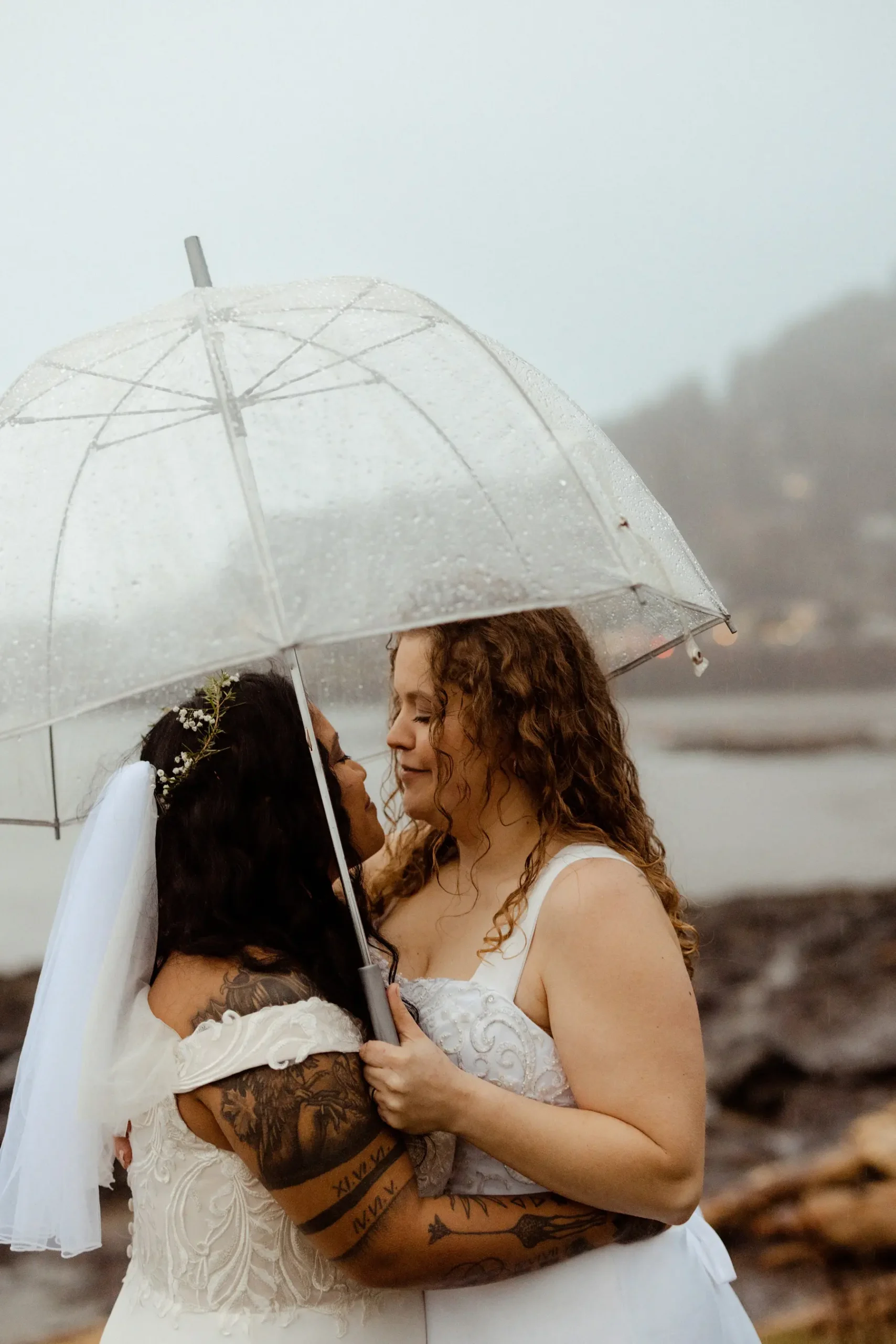 Brides Rachel and Jamie Snow embrace under a clear umbrella in the rain at Yachats Inn on the Oregon coast.