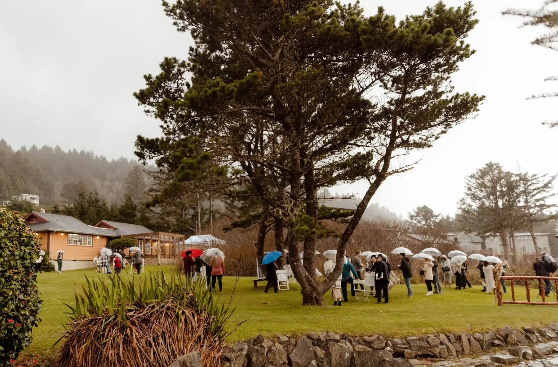 Wedding guests with umbrellas gather on a misty lawn at Yachats Inn in Oregon for Rachel and Jamie Snow's celebration.