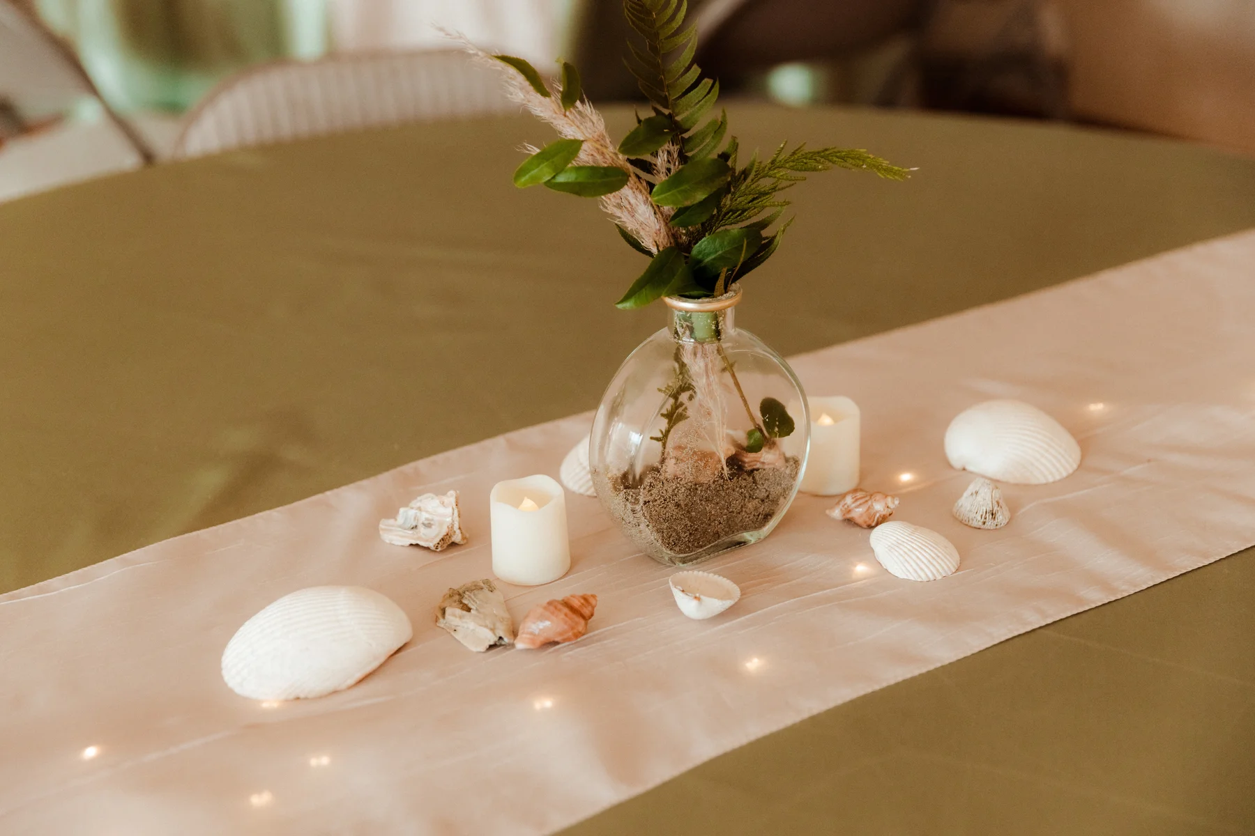 Beach-themed reception centerpiece with glass vase, sand, greenery, seashells, and LED candles at Yachats Inn.