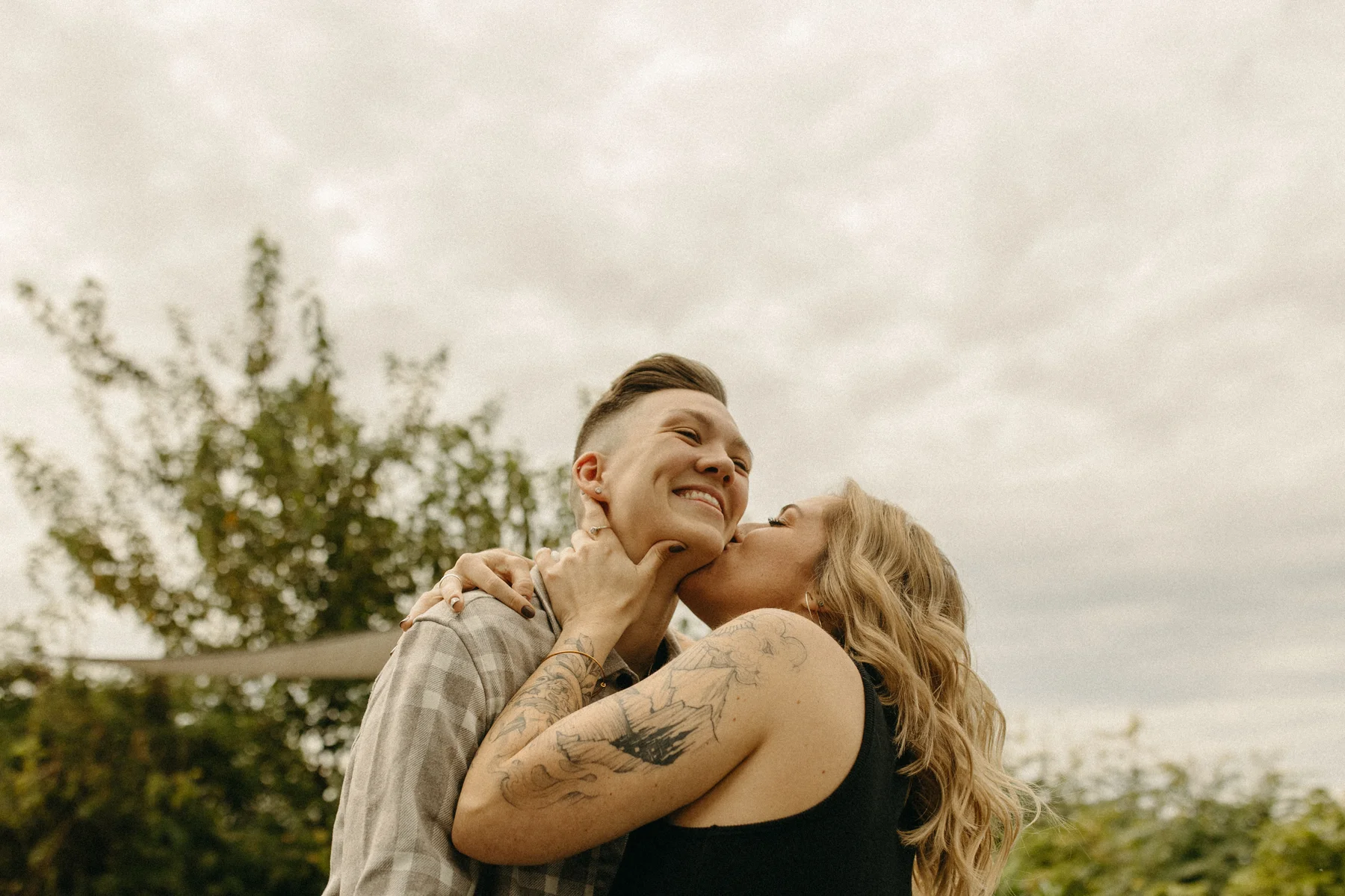 One of the partners kisses the other on the cheek as they smile during an engagement session at Gravely Brewing Co in Denver.
