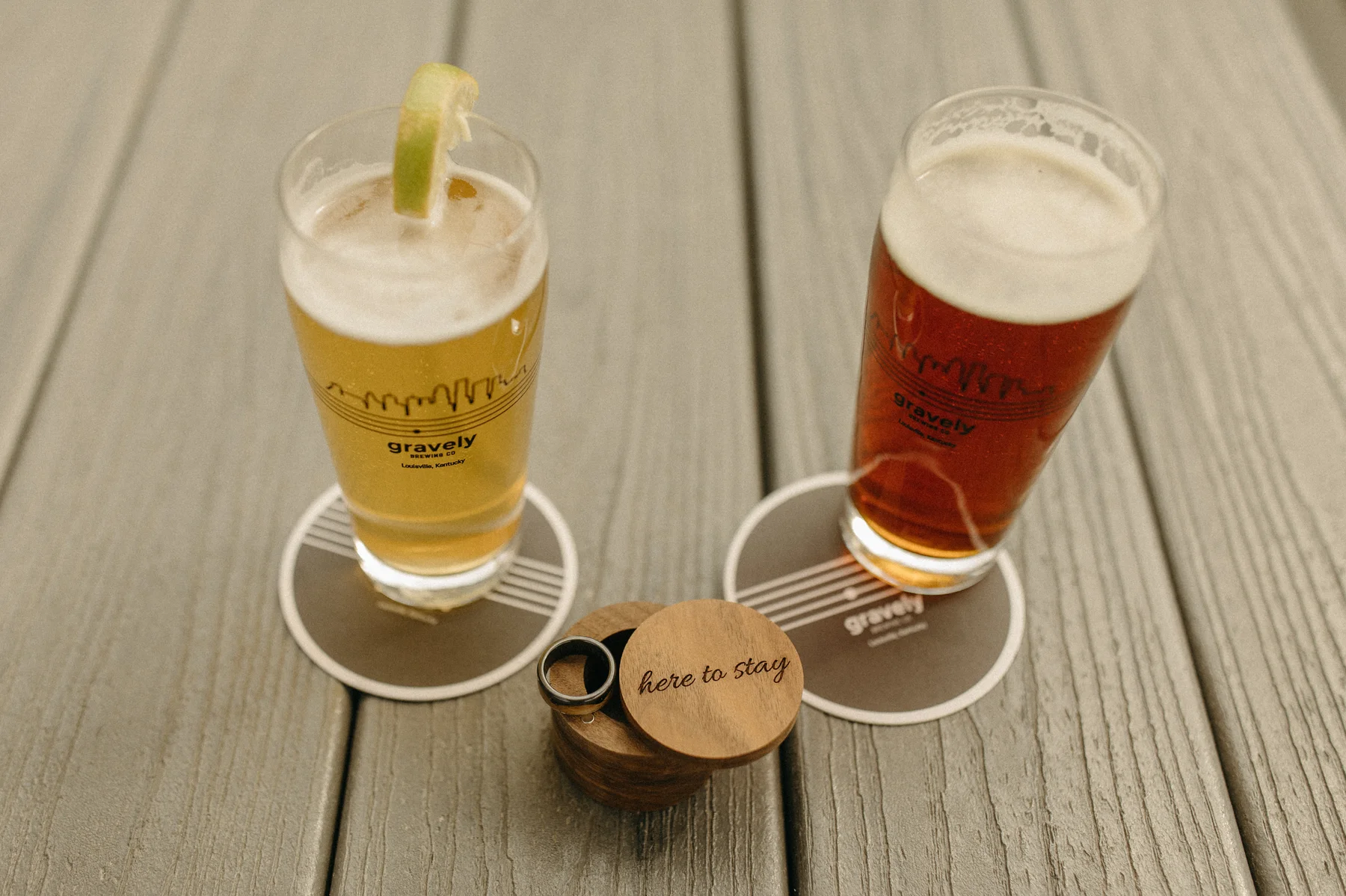 Two Gravely Brewing Co. pints and a wooden ring box engraved with 'here to stay' on a deck in Denver.
