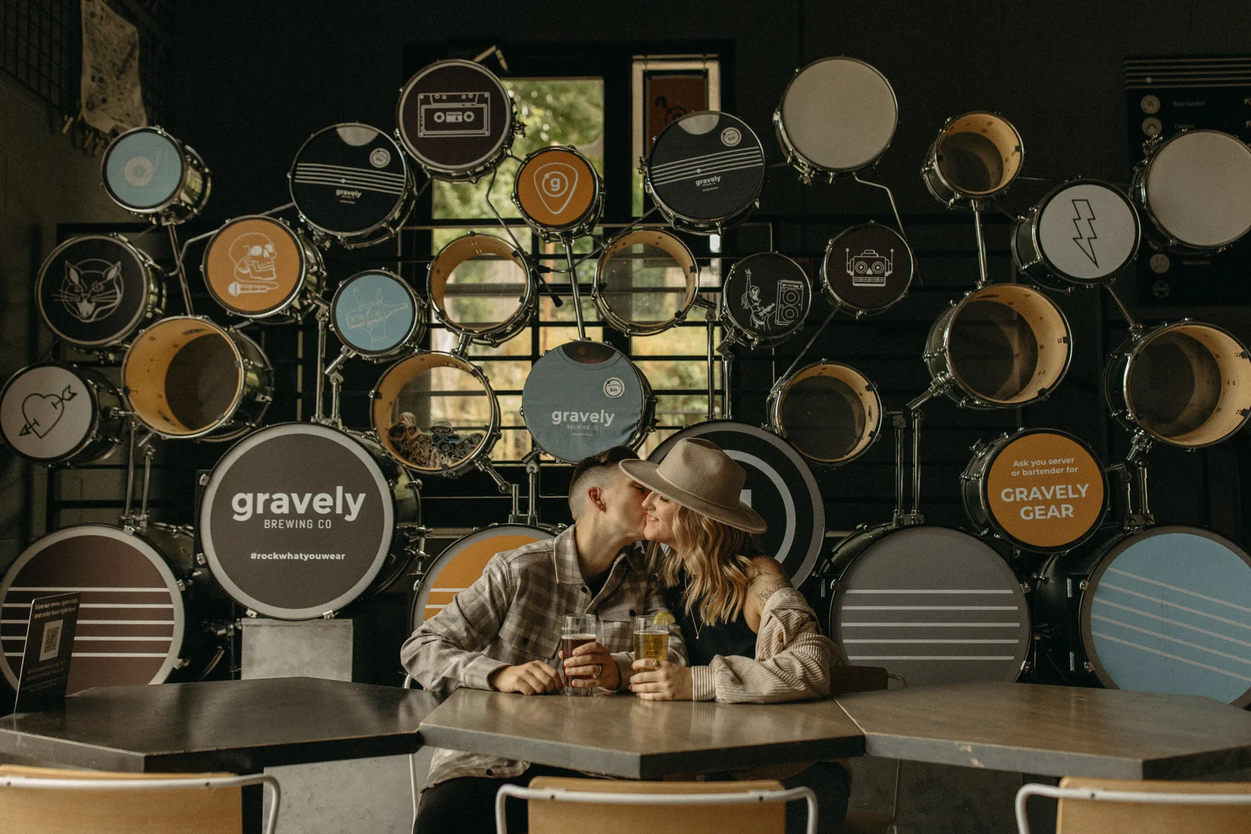 The couple shares a kiss over beers at Gravely Brewing Co in Denver, Colorado, surrounded by a wall of decorated drum heads.