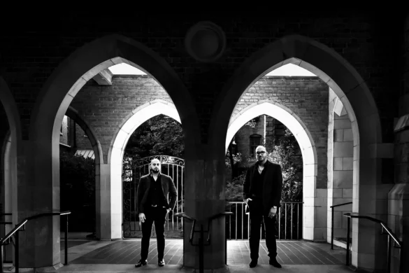 The couple poses dramatically beneath twin Gothic stone arches Raleigh, N.C., each partner framed by a separate archway in a striking black-and-white composition. Both partners are dressed in all-black attire, their confident stances contrasting with the ornate ecclesiastical architecture surrounding them. Photographed by Kivus and Camera, this moody engagement session beautifully captures the couple's bold aesthetic against the historic brick and stonework of the venue.