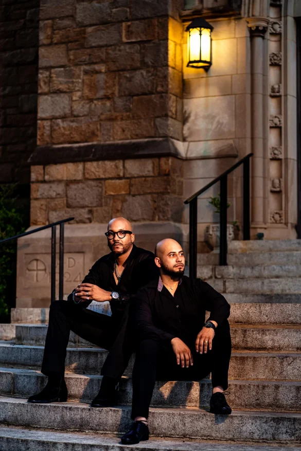 Two partners sit back-to-back on stone steps, both dressed in all black under a warm lantern glow.