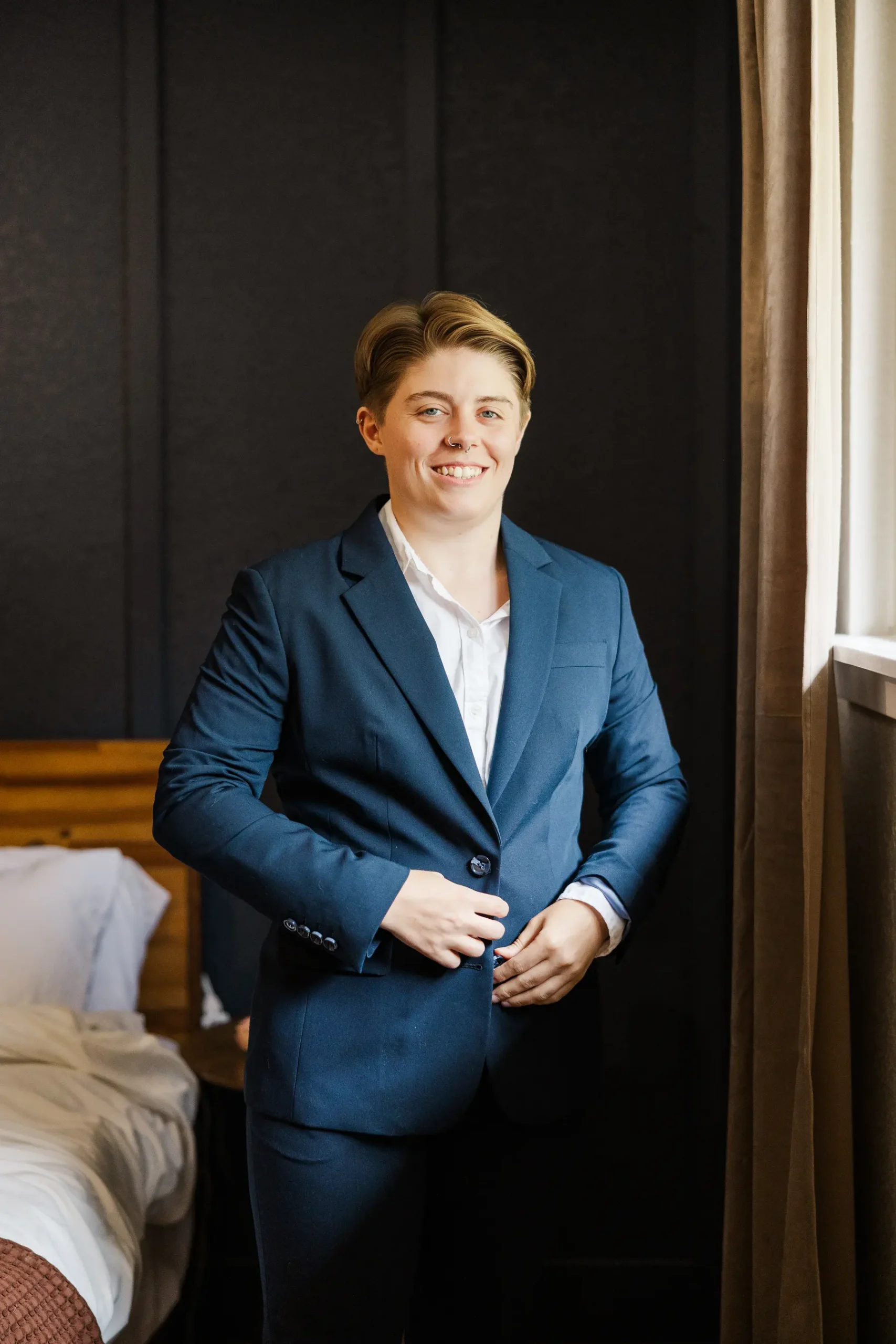 A person in a navy blue suit buttons their jacket while smiling in a hotel room at Halfway House in Boulder, CO.