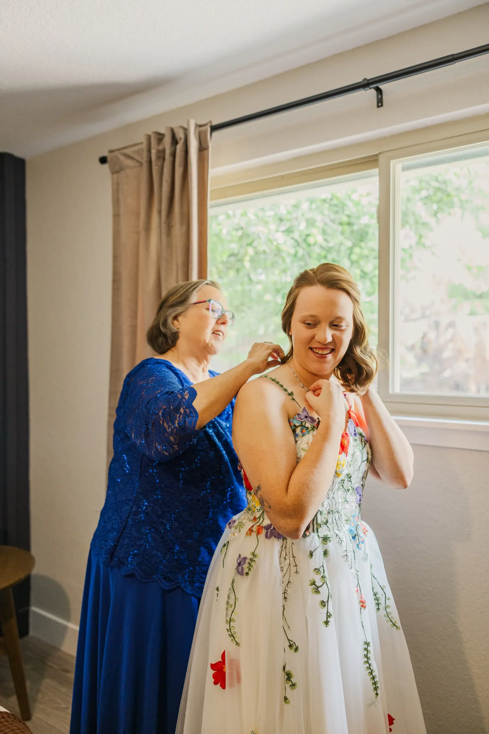 A woman in a blue lace dress helps bride Hannah Rusow with her necklace as she gets ready in Boulder, CO.