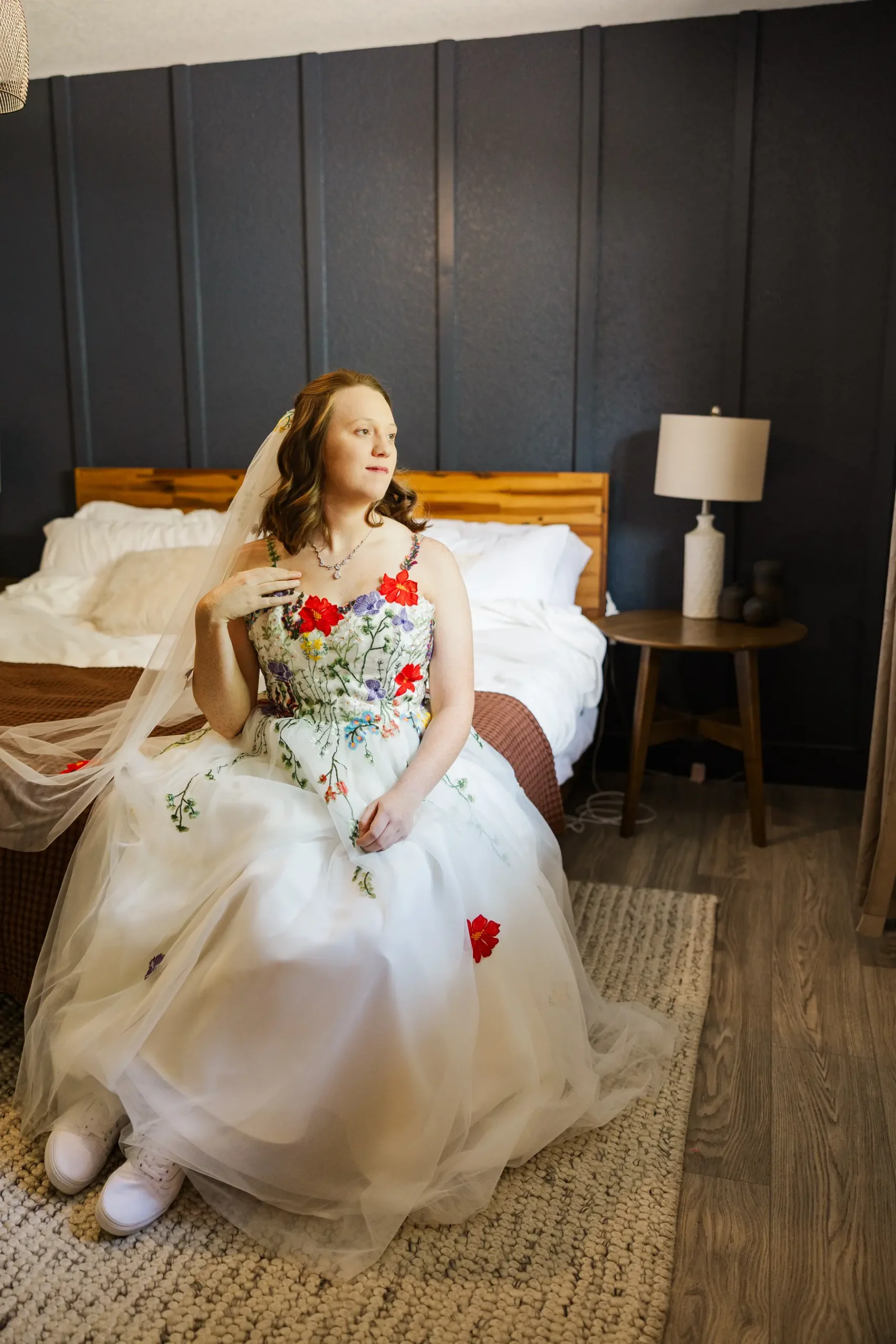 Bride Sydney Rusow sits on a bed in a floral embroidered wedding gown and veil at Halfway House in Boulder, CO.