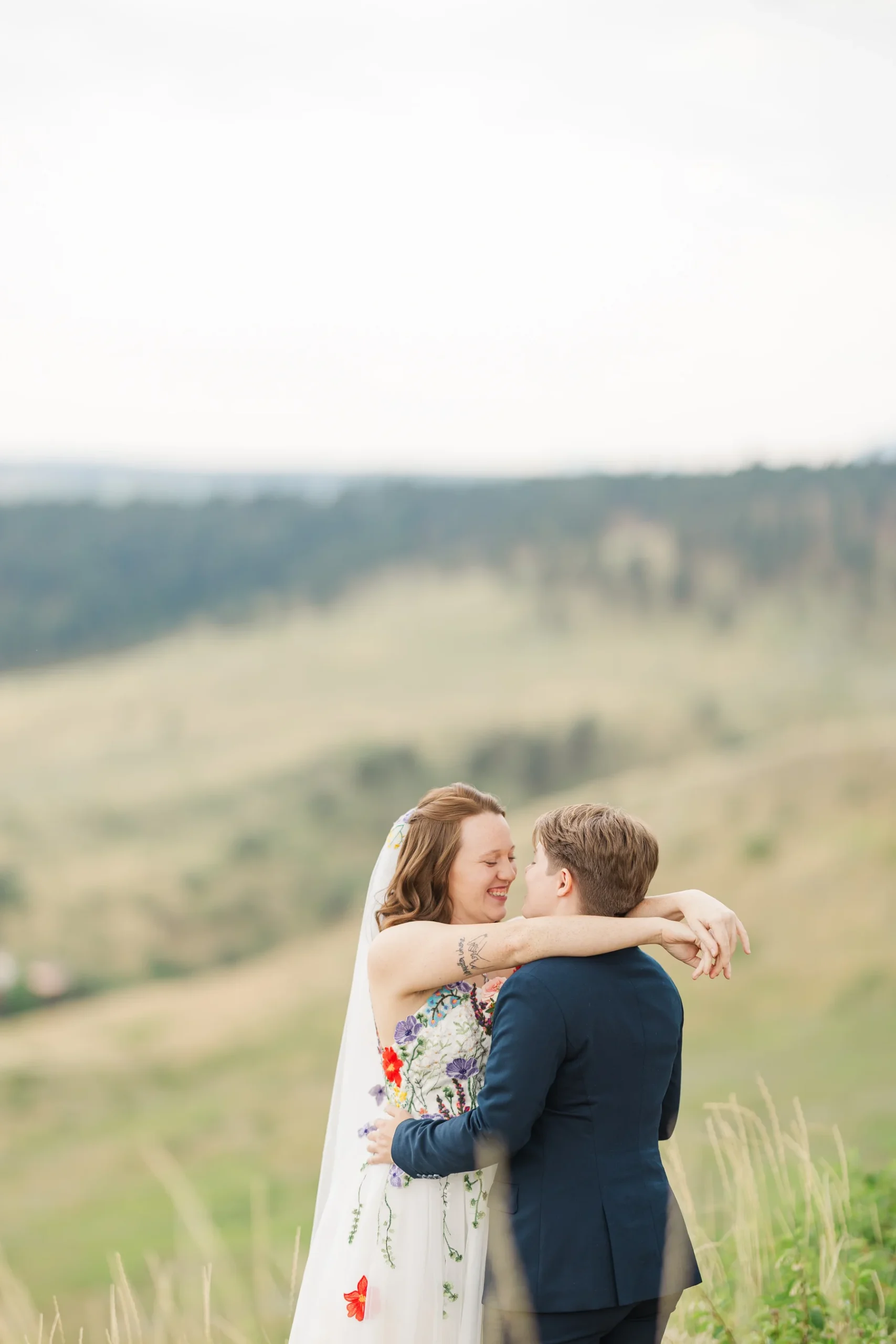 Sydney and Hannah Rusow embrace and smile at each other on a grassy hillside at Halfway House in Boulder, CO.