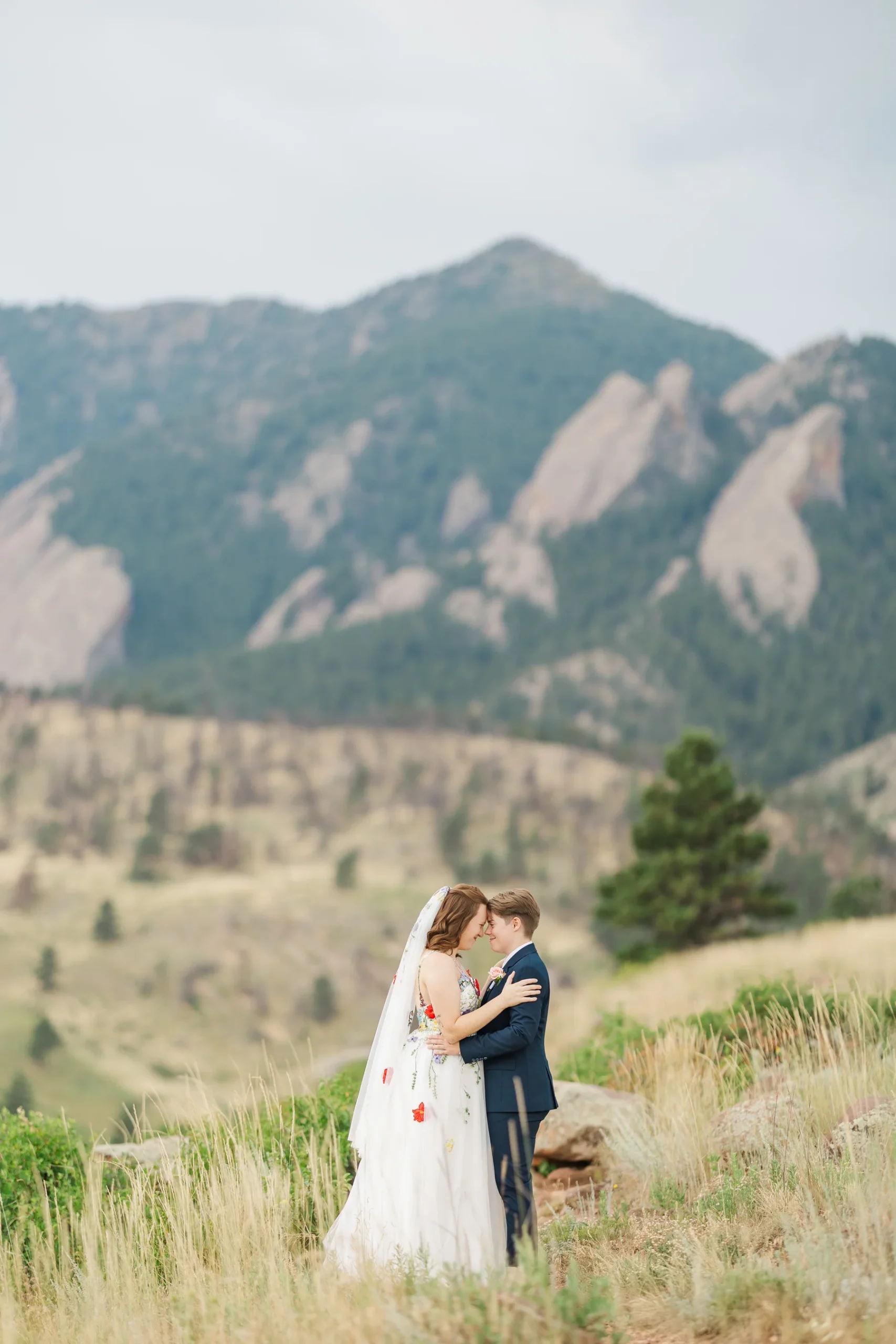 Sydney and Hannah Rusow share an intimate moment in a floral wedding gown and navy suit with the Boulder Flatirons behind them.