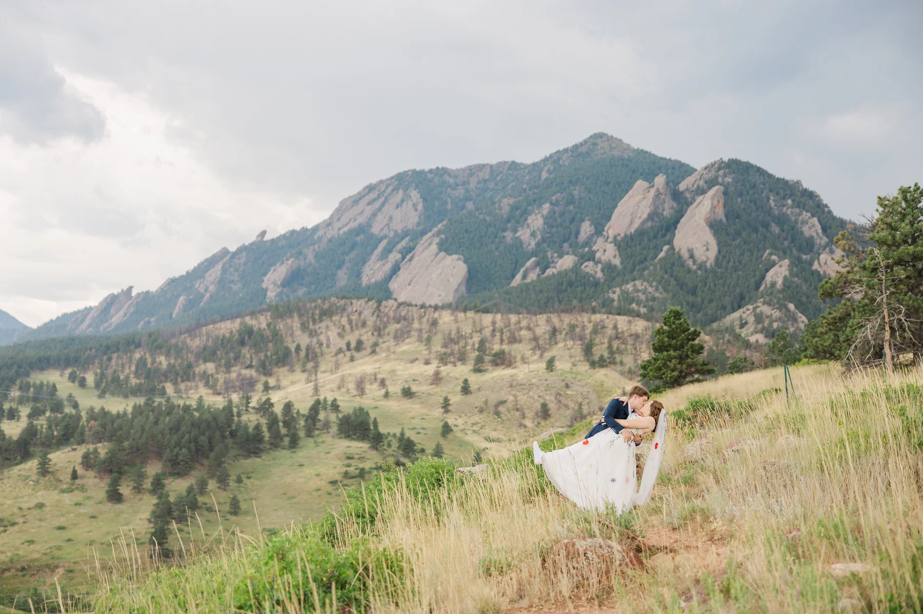 Sydney and Hannah Rusow share a romantic dip kiss on a hillside with the Flatirons of Boulder, CO, behind them.