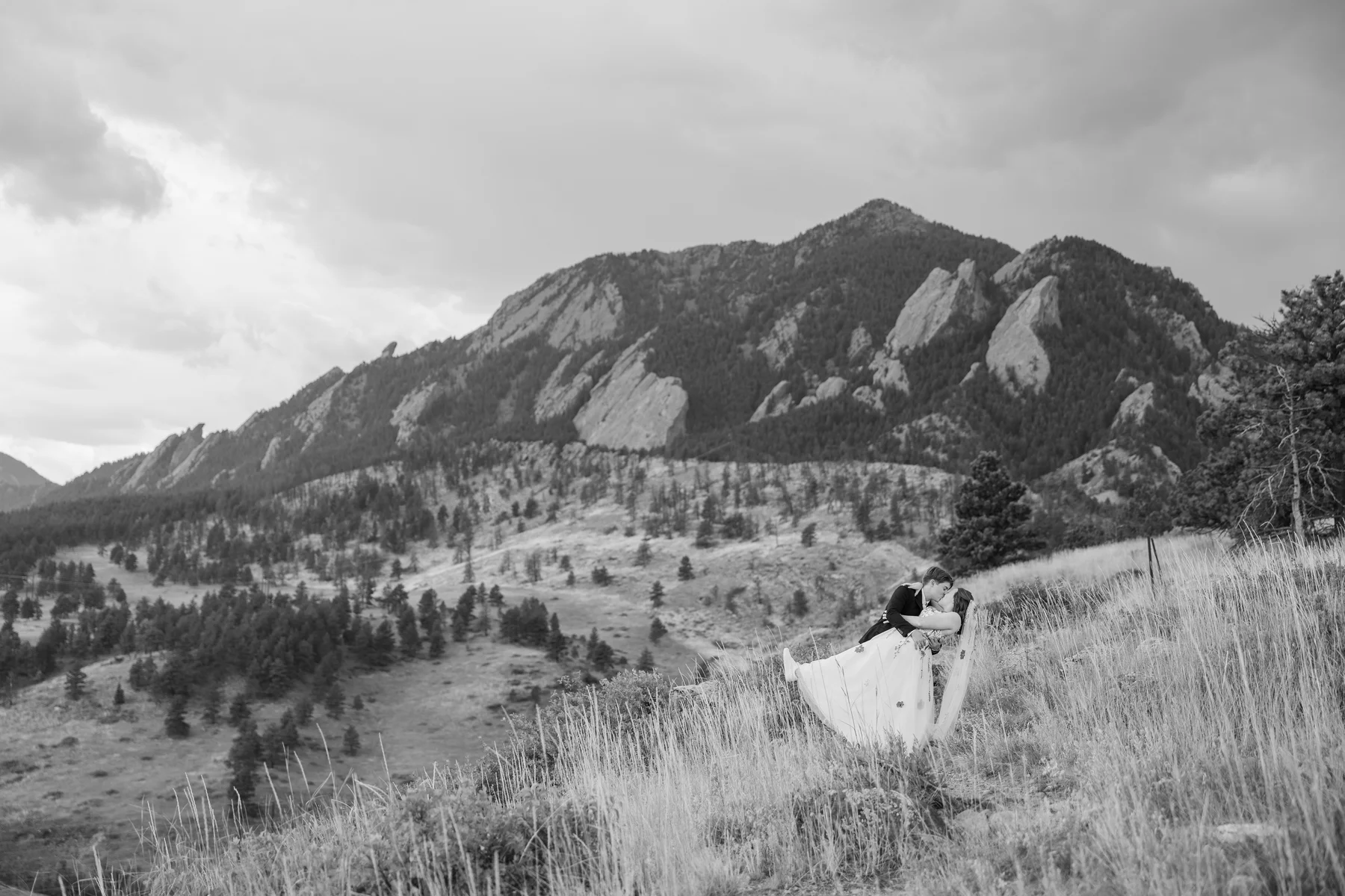 Sydney and Hannah Rusow share a romantic dip kiss on a hillside with the Flatirons of Boulder, CO, behind them.
