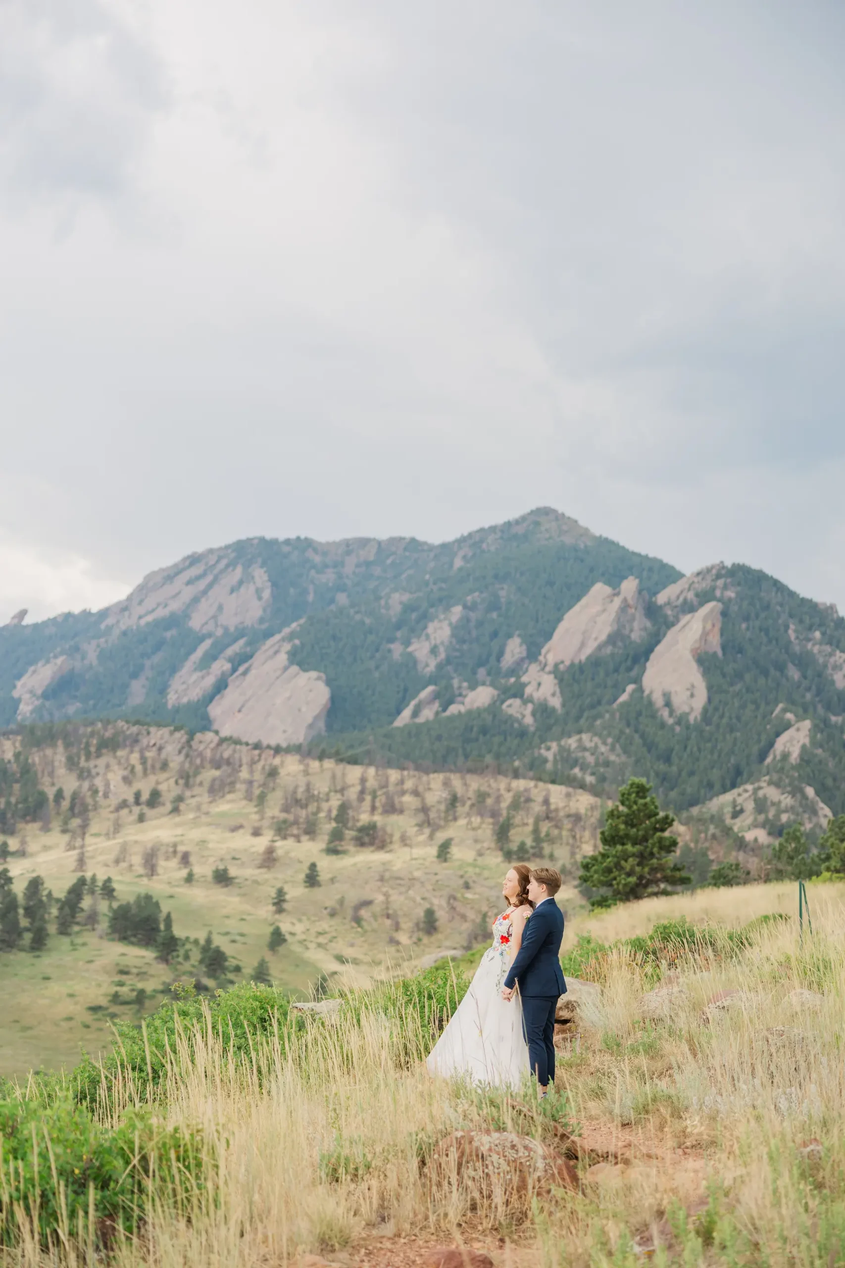 Sydney and Hannah Rusow stand together in a white floral gown and navy suit with the Boulder Flatirons behind them.