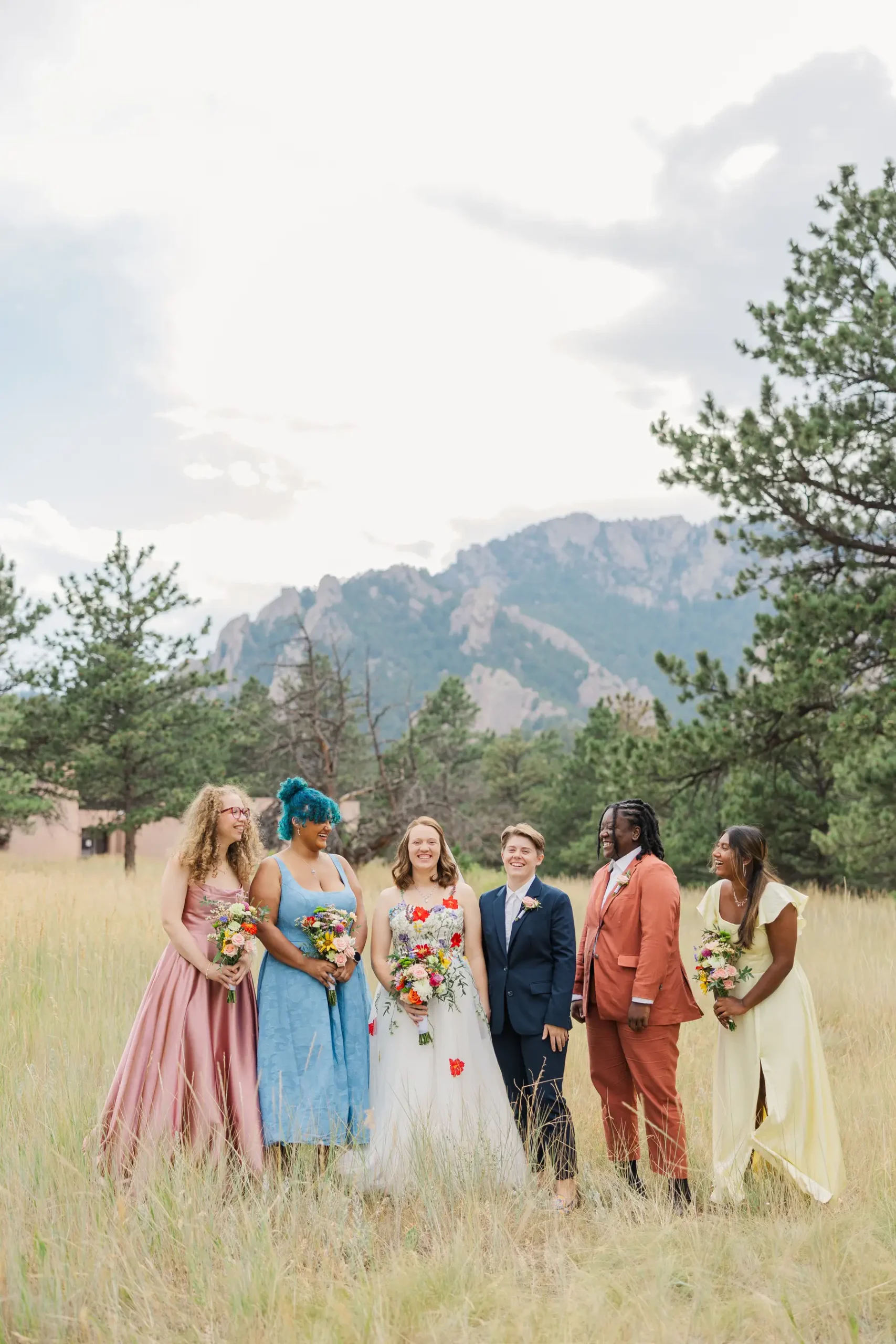 Sydney and Hannah Rusow laugh with their wedding party in a mountain meadow at Halfway House in Boulder, CO.