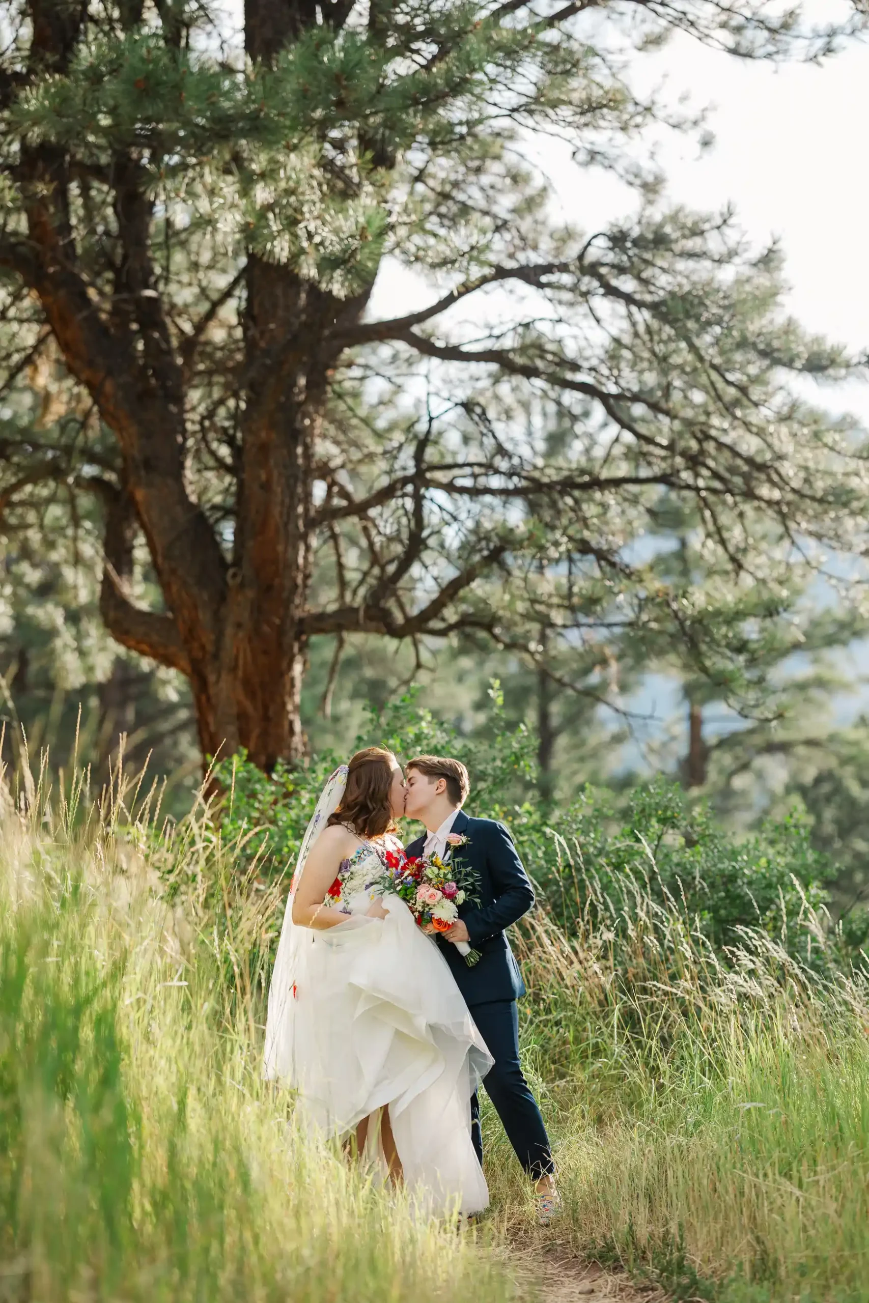 Sydney and Hannah Rusow share a kiss in tall grass beneath a pine tree at Halfway House in Boulder, CO.