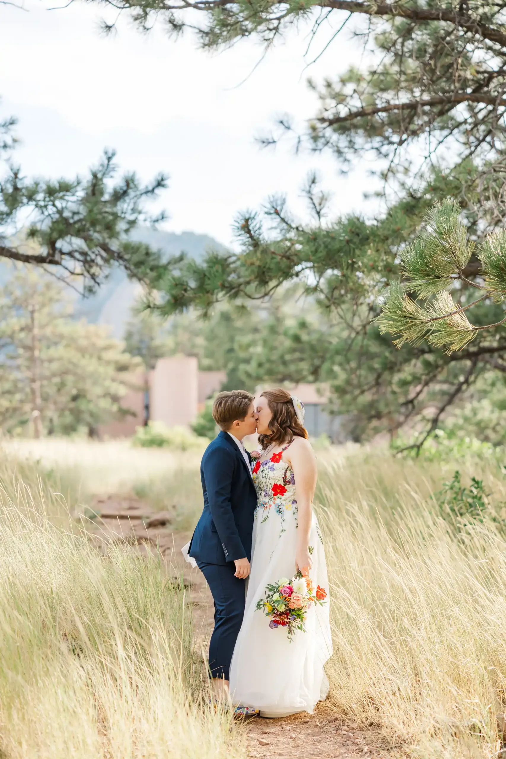 Sydney and Hannah Rusow share a kiss on a nature trail at Halfway House in Boulder, CO, surrounded by golden grasses and pine trees.