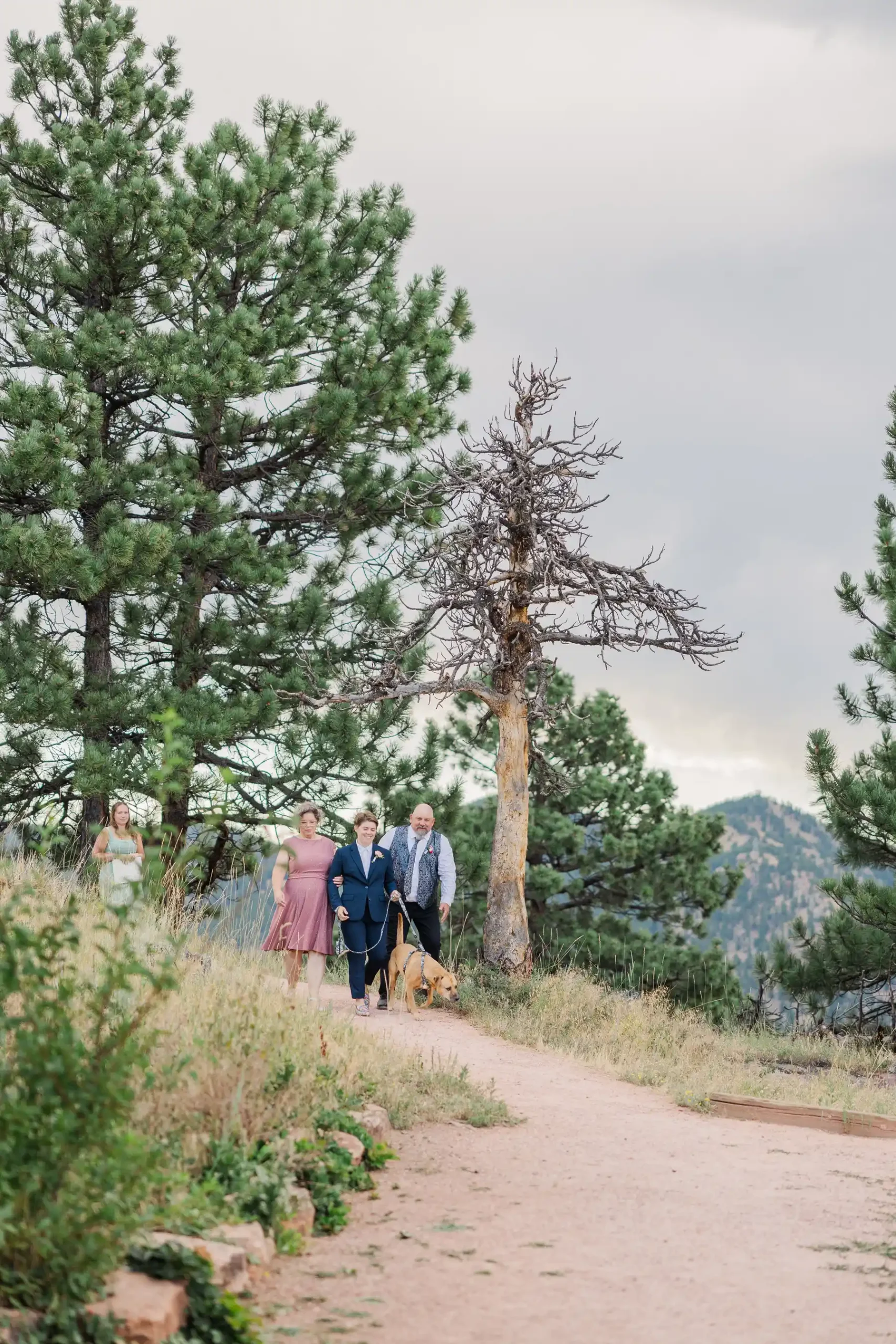 Sydney or Hannah Rusow walks down a pine-lined dirt path with two escorts and a dog at Halfway House in Boulder, CO.