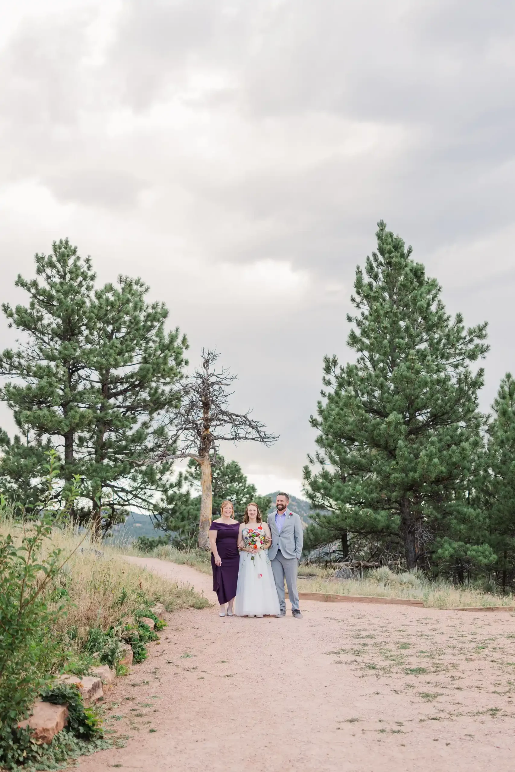 Sydney and Hannah Rusow pose with a guest on a dirt path surrounded by pine trees at Halfway House in Boulder, CO.