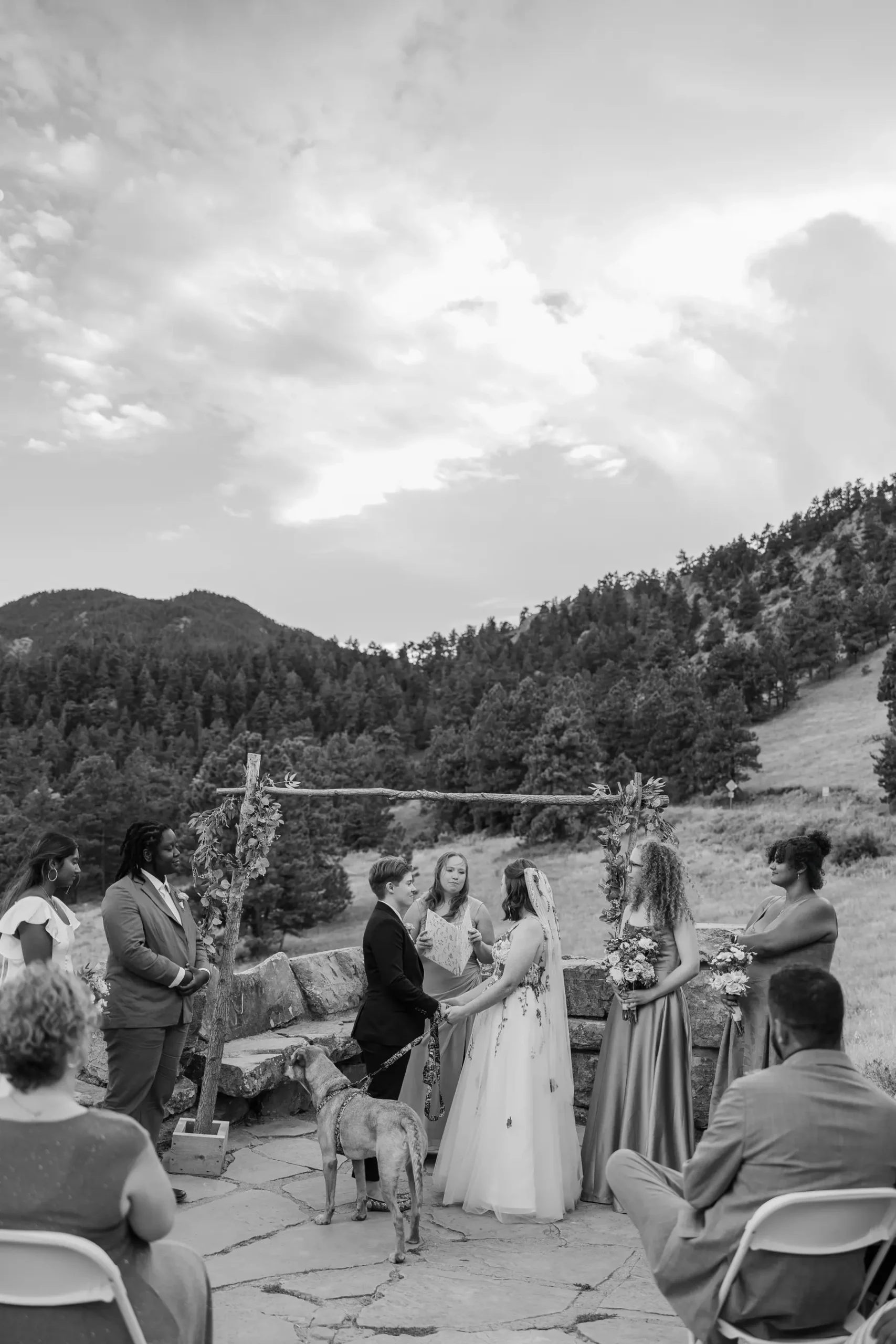 Sydney and Hannah Rusow exchange vows under a rustic floral arch at Halfway House in Boulder, CO, with mountains behind.