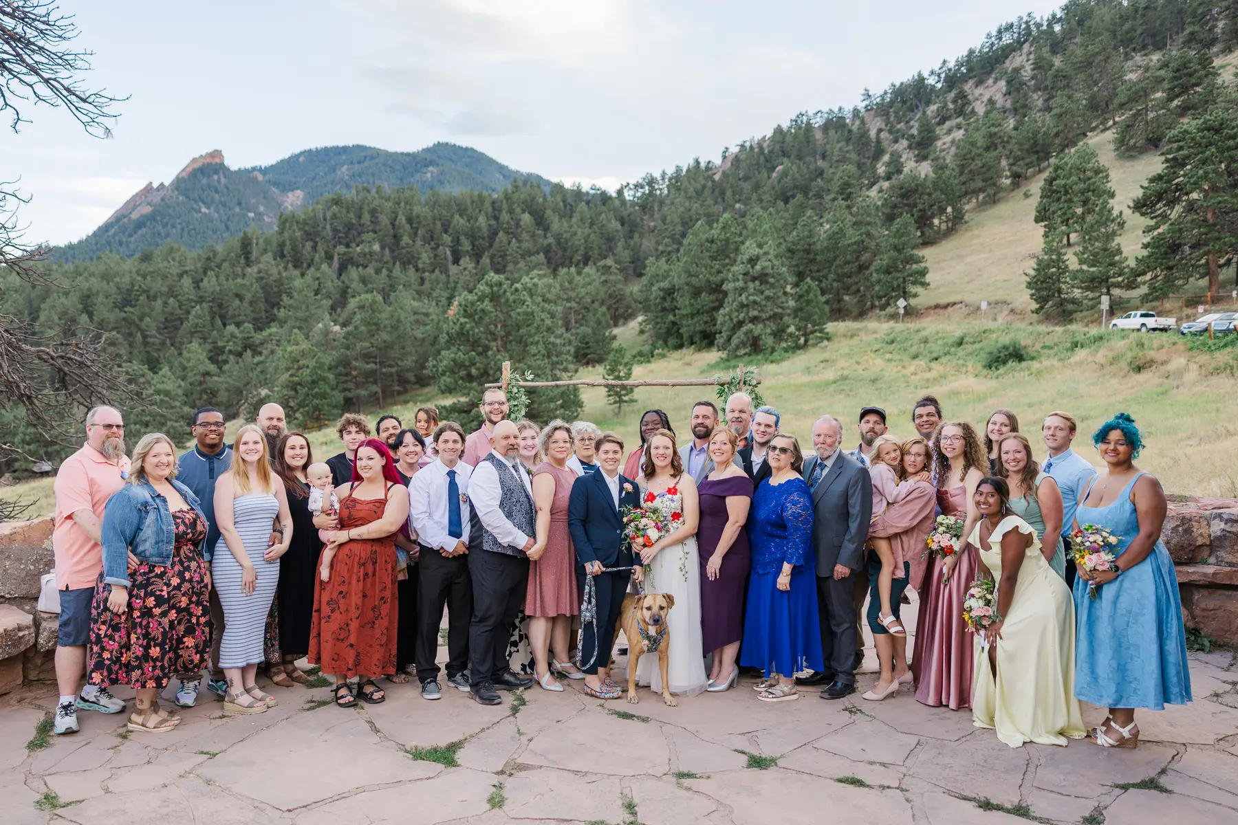 Sydney and Hannah Rusow pose with wedding guests and their dog at Halfway House in Boulder, CO, with the Flatirons in the background.