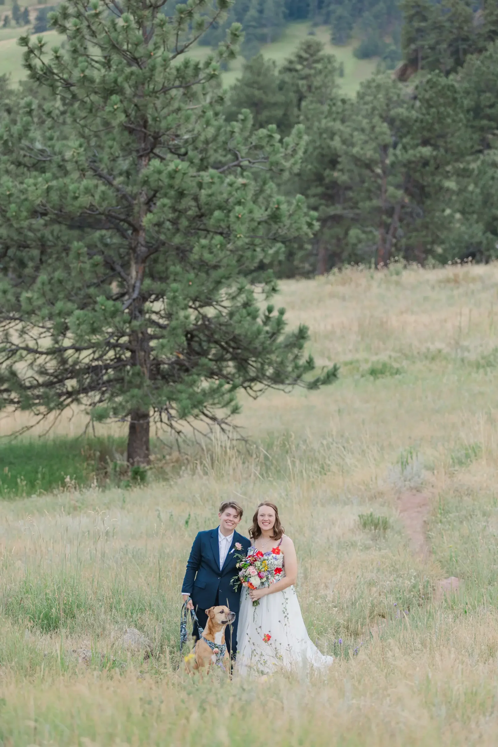 Sydney and Hannah Rusow smile in a wildflower meadow with their dog at Halfway House in Boulder, CO.