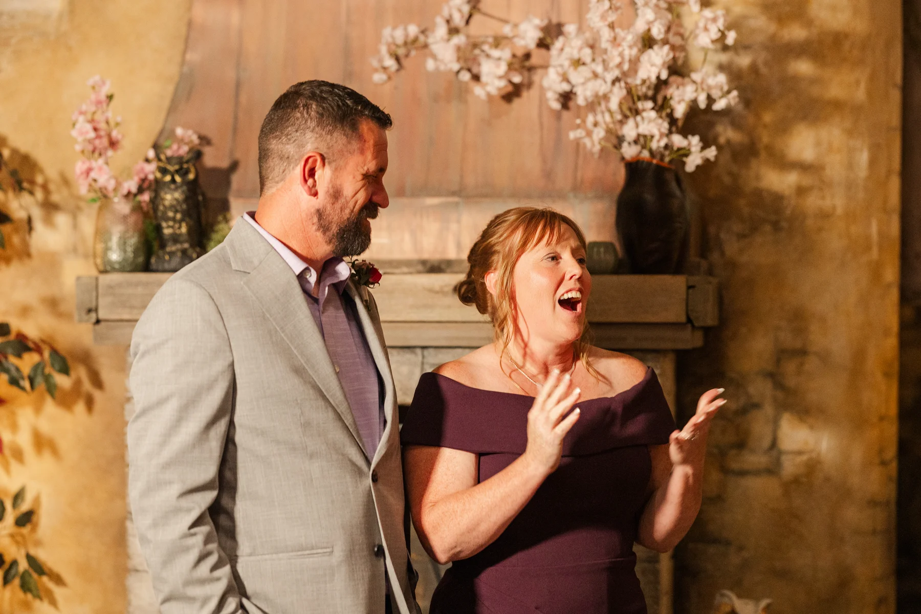 A man in a gray suit and woman in a plum off-shoulder dress laugh together at the Halfway House reception in Boulder, CO.