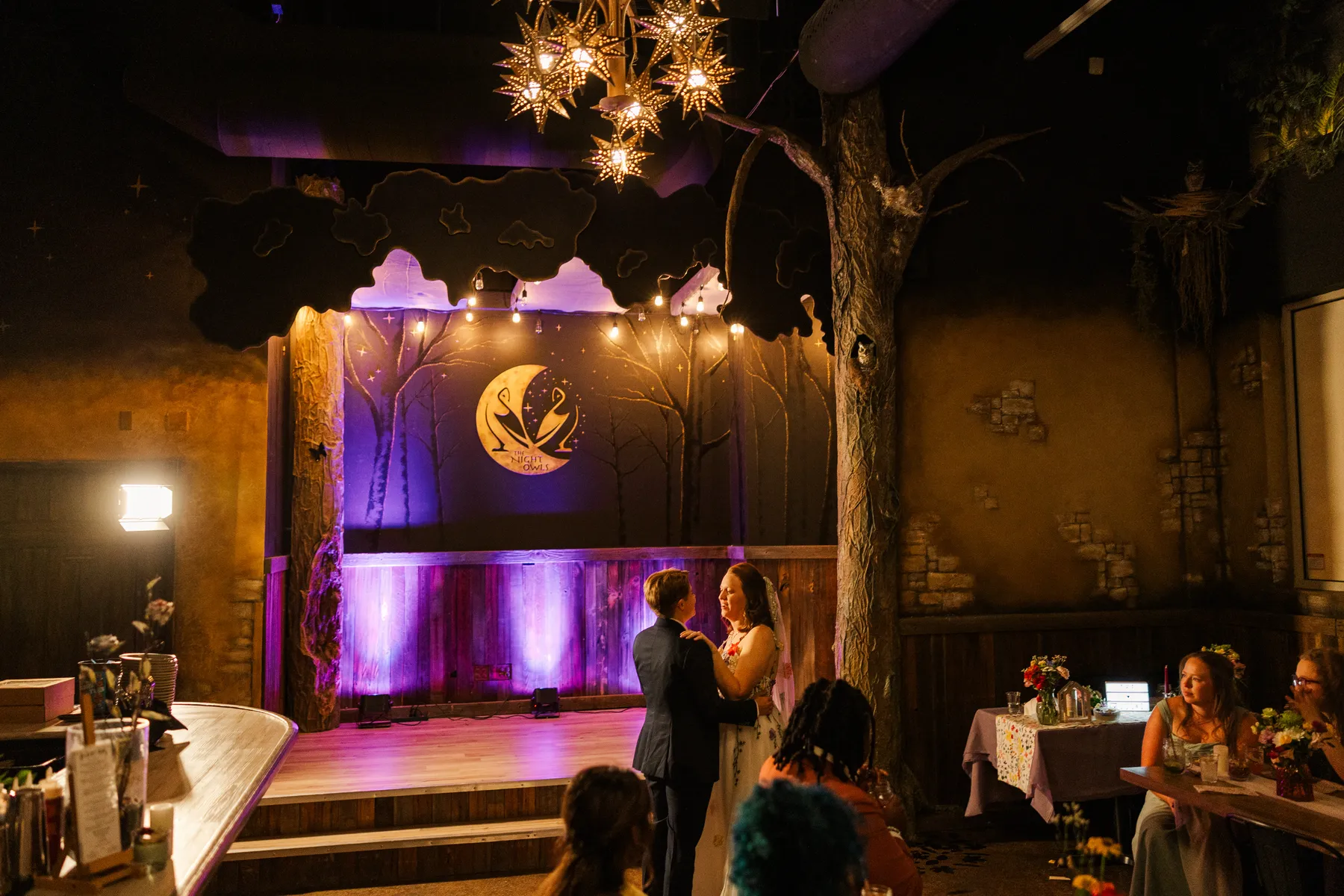 Sydney and Hannah Rusow share their first dance at Halfway House in Boulder, CO, beneath star chandeliers and purple stage lighting.