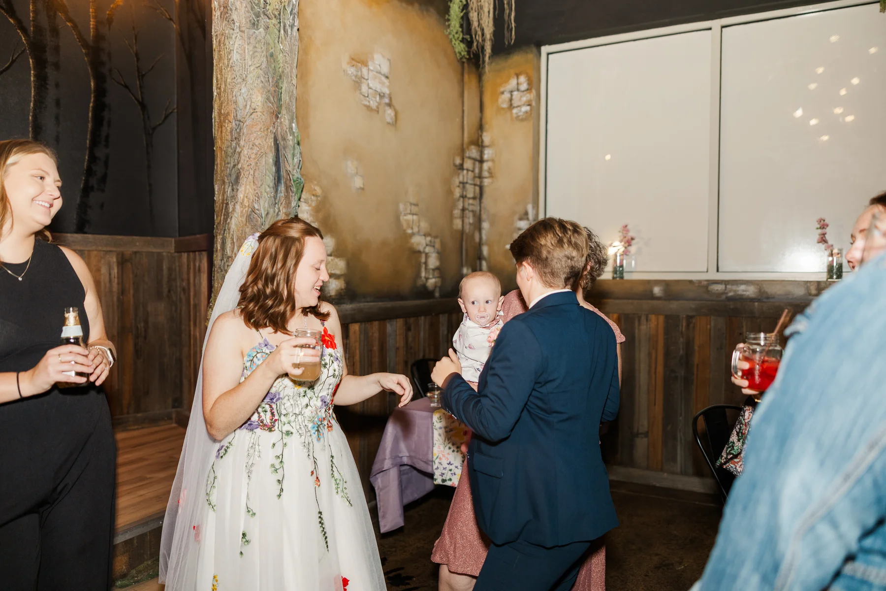 Bride Sydney Rusow in floral gown laughs while dancing with guests including a baby at Halfway House reception in Boulder, CO.