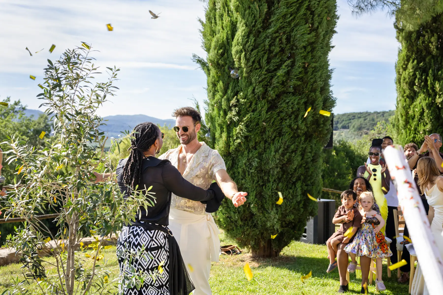 Two grooms walk a confetti-lined aisle outdoors at Conti di San Bonifacio Wine Resort in Tuscany, Italy, as guests cheer.
