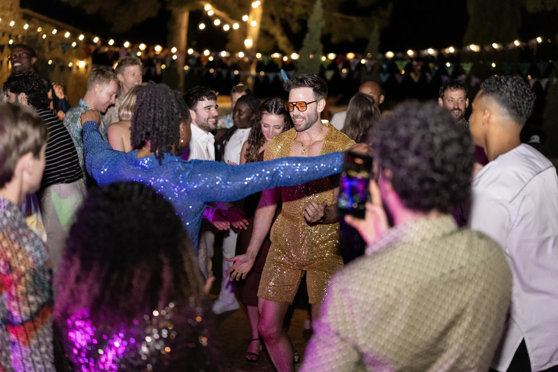 A groom in a gold sequin outfit dances joyfully with guests at an outdoor evening reception at Conti di San Bonifacio, Tuscany.