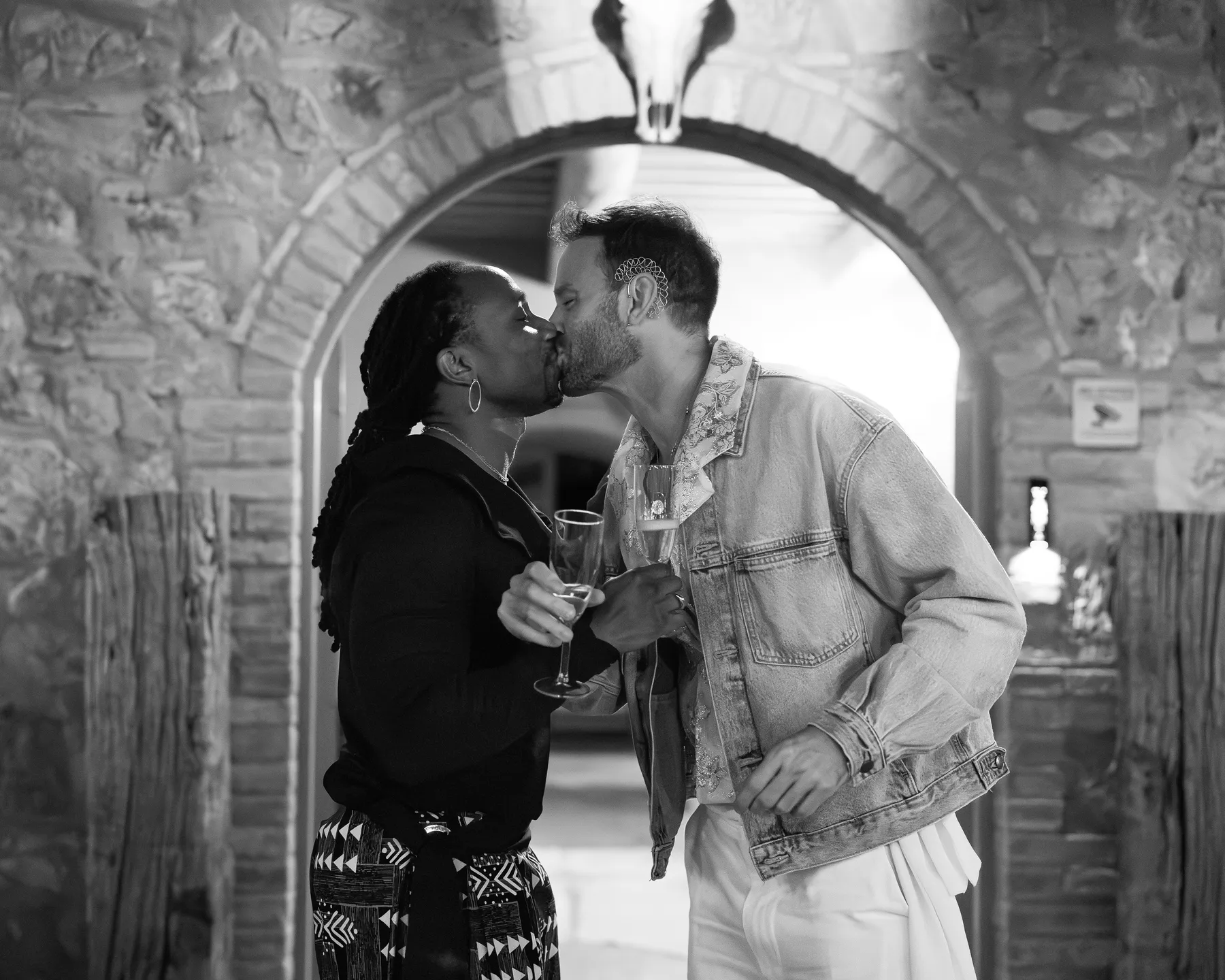 Two grooms share a kiss while holding champagne flutes beneath a stone archway at Conti di San Bonifacio Wine Resort in Tuscany.