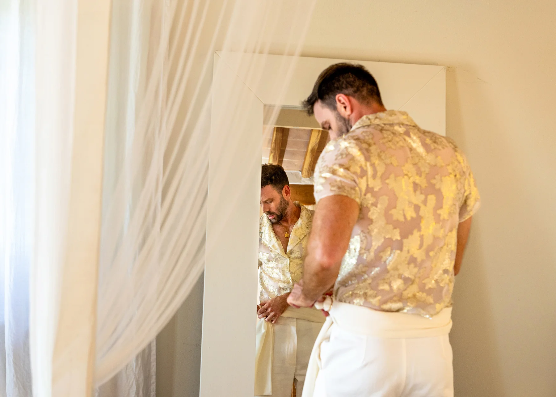 A groom in a gold brocade shirt adjusts his white trousers while checking his reflection in a mirror during pre-ceremony prep.