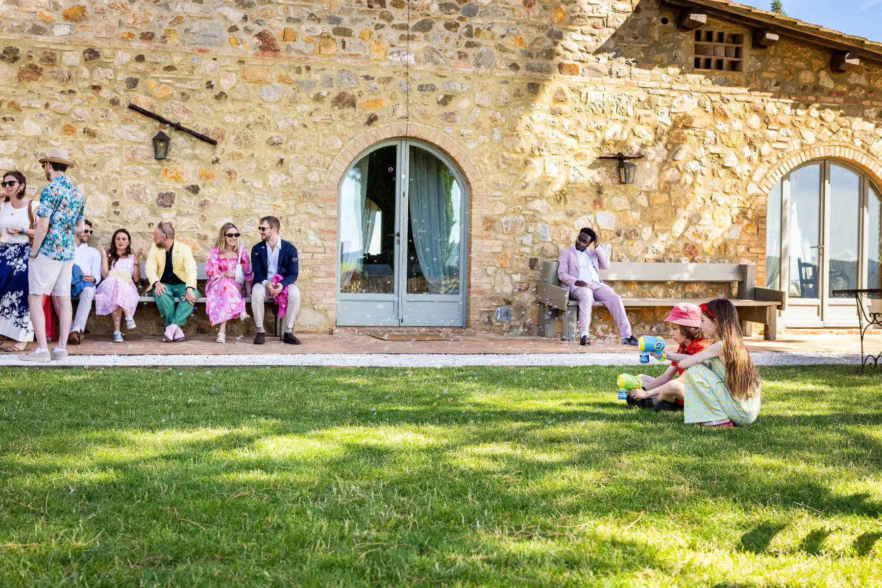 Wedding guests relax on benches outside a stone villa while two children play with bubble guns on the lawn at Conti di San Bonifacio.