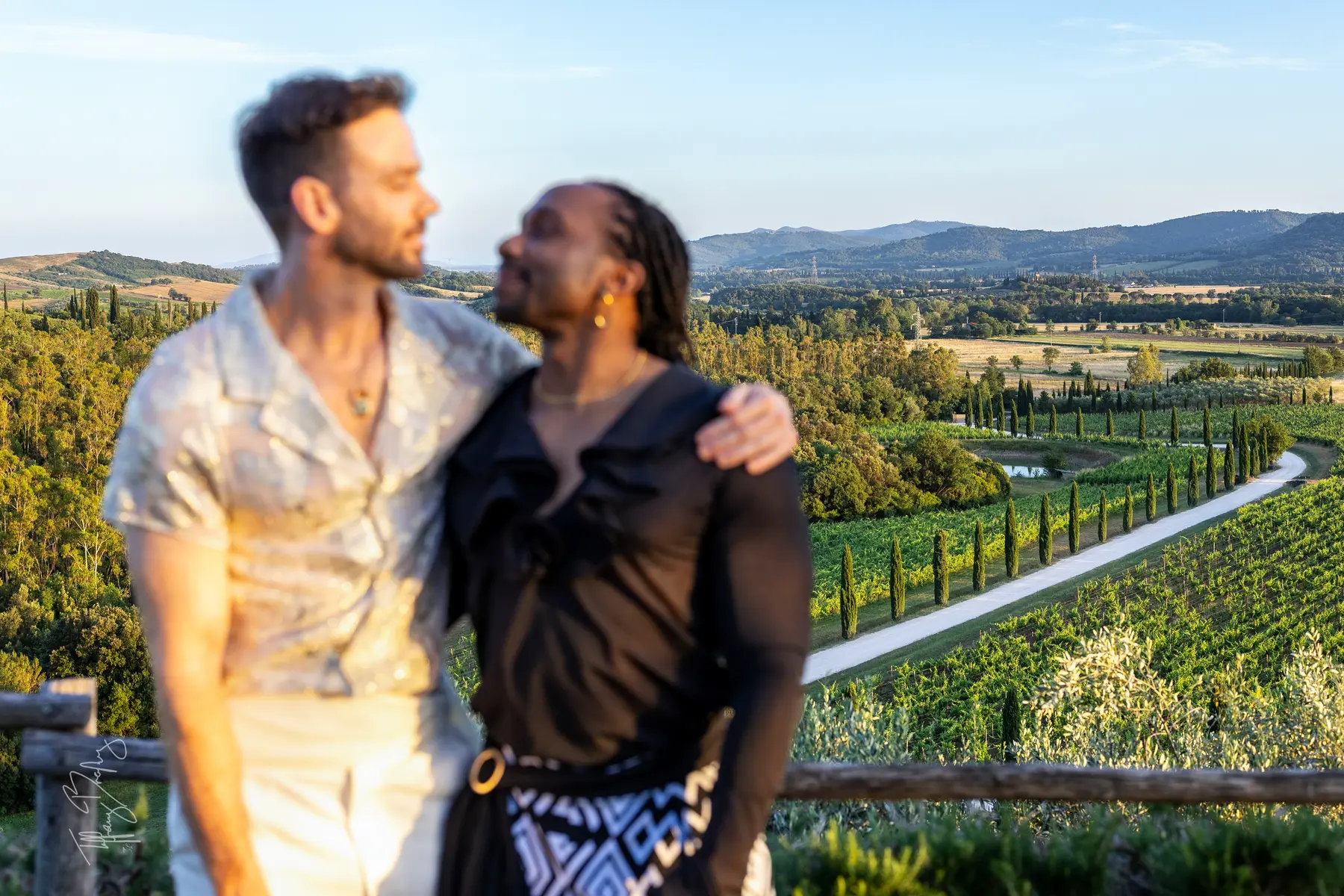 Two grooms embrace on a terrace overlooking the cypress-lined vineyards and rolling hills of Conti di San Bonifacio Wine Resort in Tuscany.