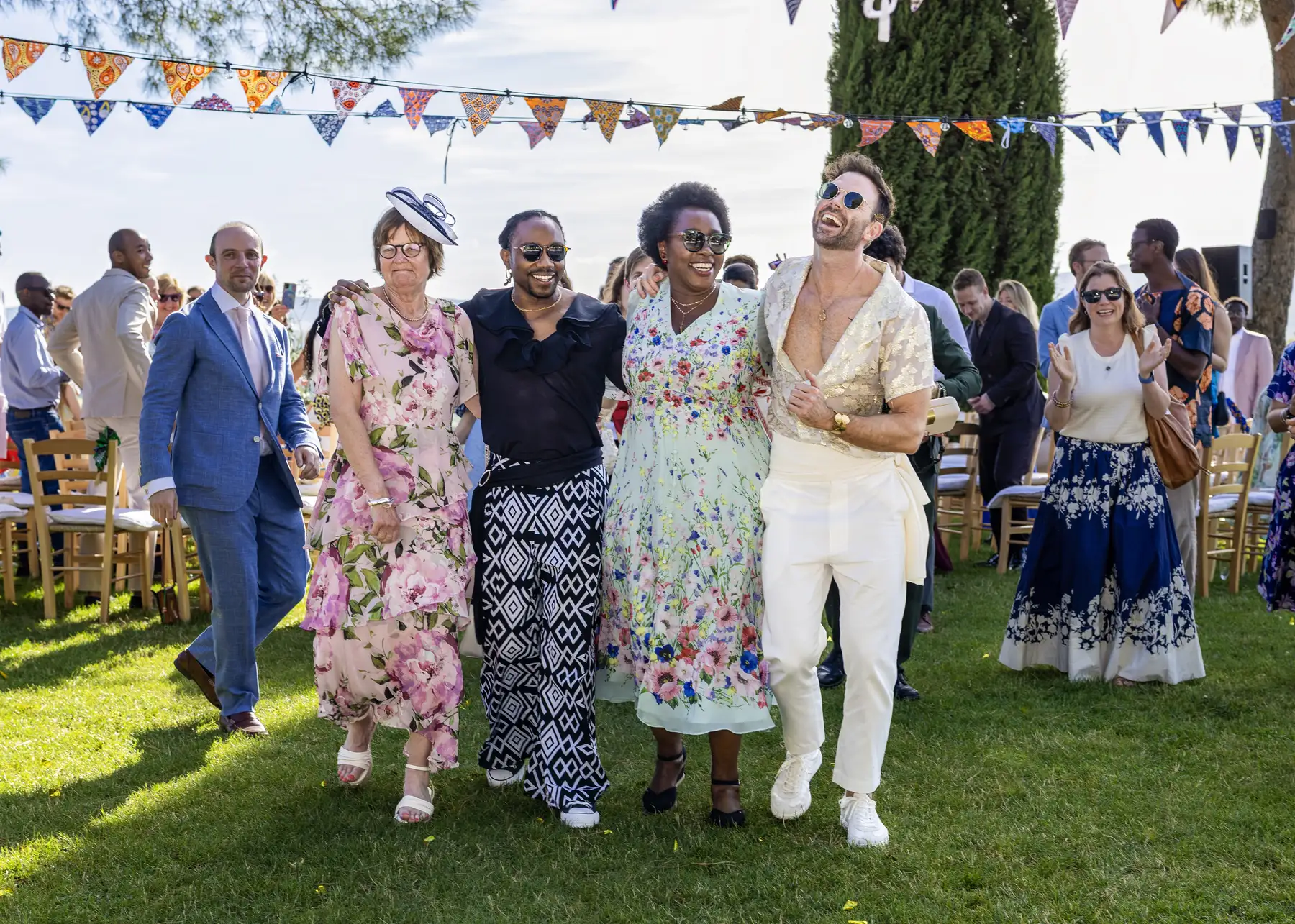 Two grooms walk arm-in-arm with guests on the lawn at Conti di San Bonifacio wine resort in Tuscany, laughing joyfully.