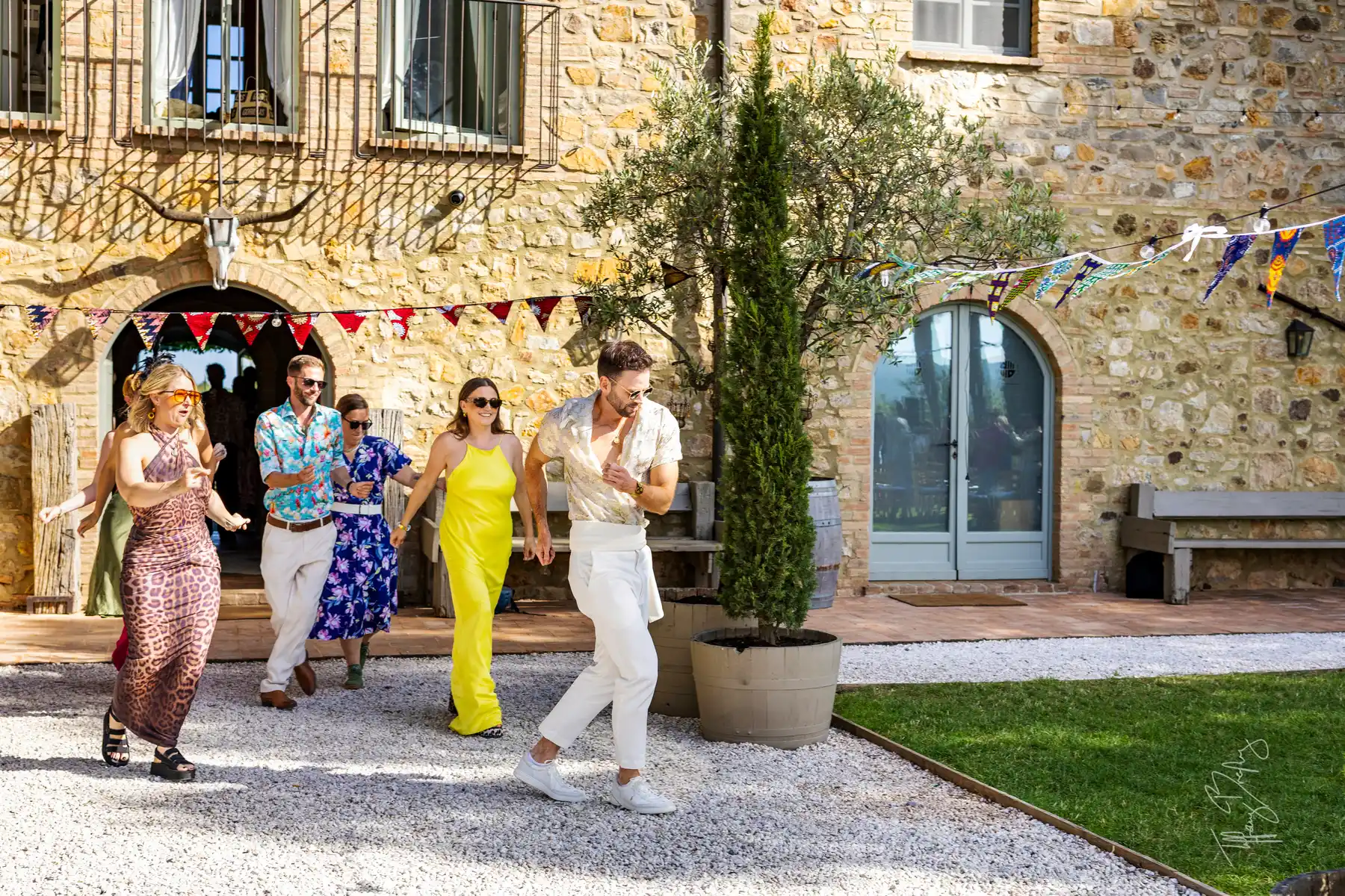 Wedding guests in colorful summer attire walk through the sun-drenched courtyard at Conti di San Bonifacio Wine Resort in Tuscany.