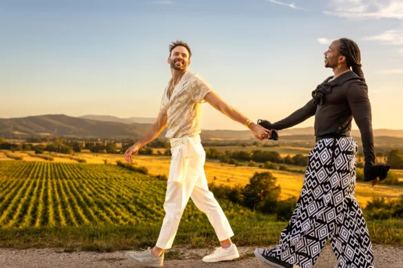 Two grooms walk hand in hand along a sunlit road overlooking vineyards at Conti di San Bonifacio Wine Resort in Tuscany.