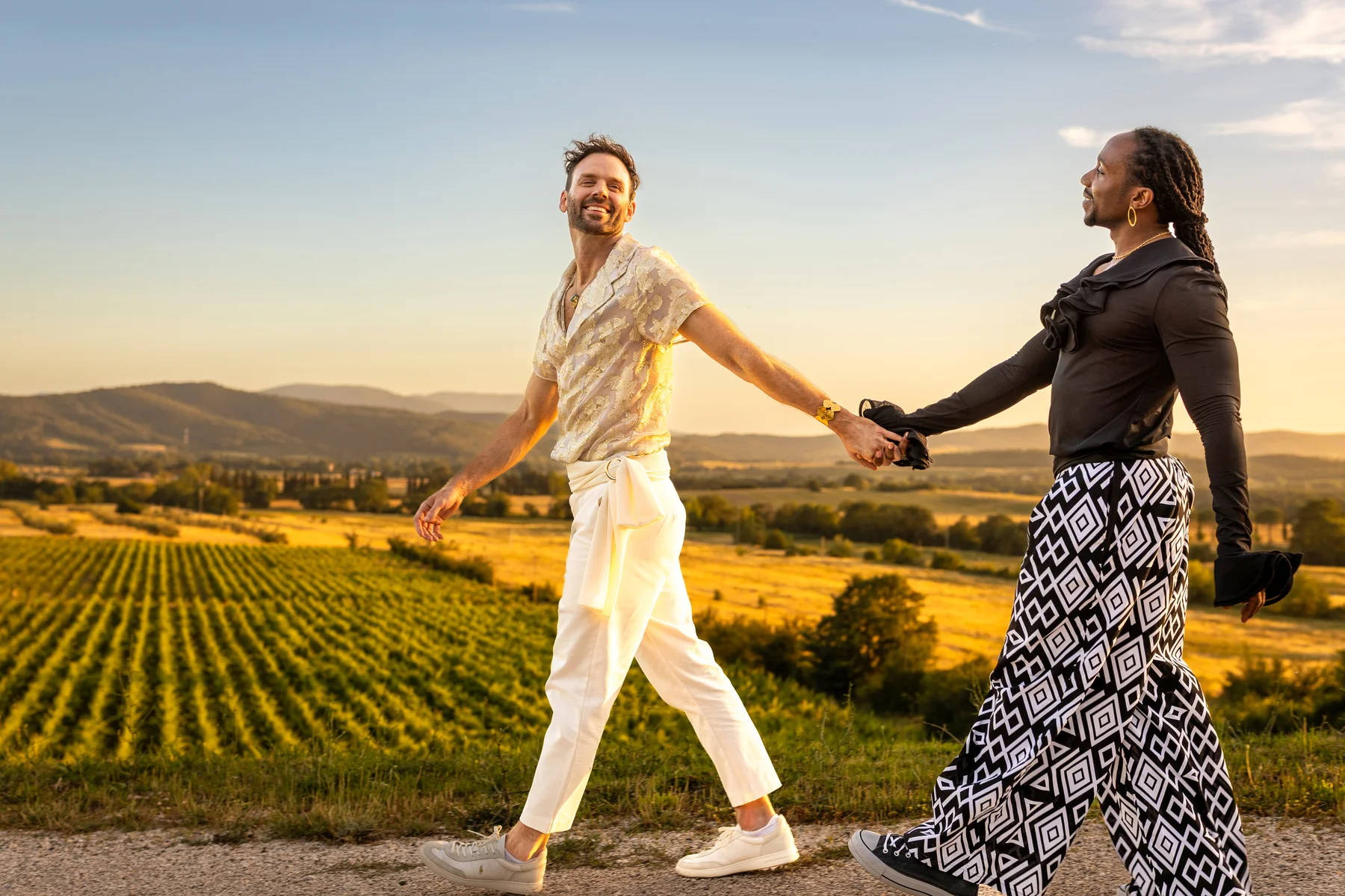 Two grooms walk hand in hand along a sunlit road overlooking vineyards at Conti di San Bonifacio Wine Resort in Tuscany.