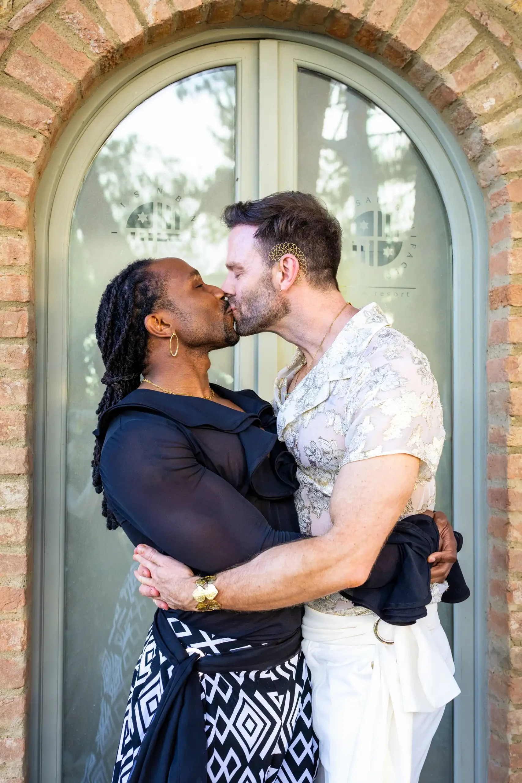 Two grooms share a kiss outside an arched brick doorway at Conti di San Bonifacio Wine Resort in Tuscany, Italy.