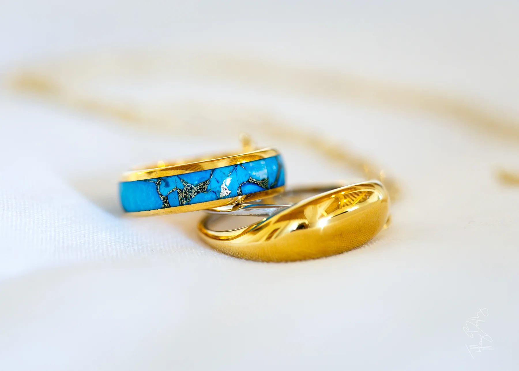 Two gold wedding bands โ one with a turquoise inlay, one a polished dome โ rest on white fabric at Conti di San Bonifacio.
