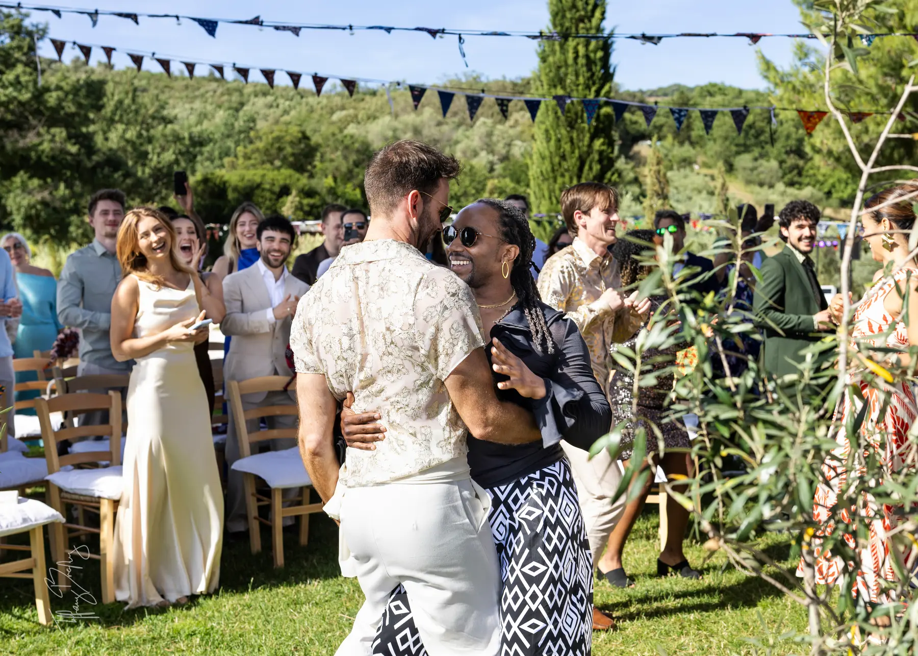 Two grooms share a joyful dance outdoors at Conti di San Bonifacio Wine Resort in Tuscany as smiling guests cheer around them.