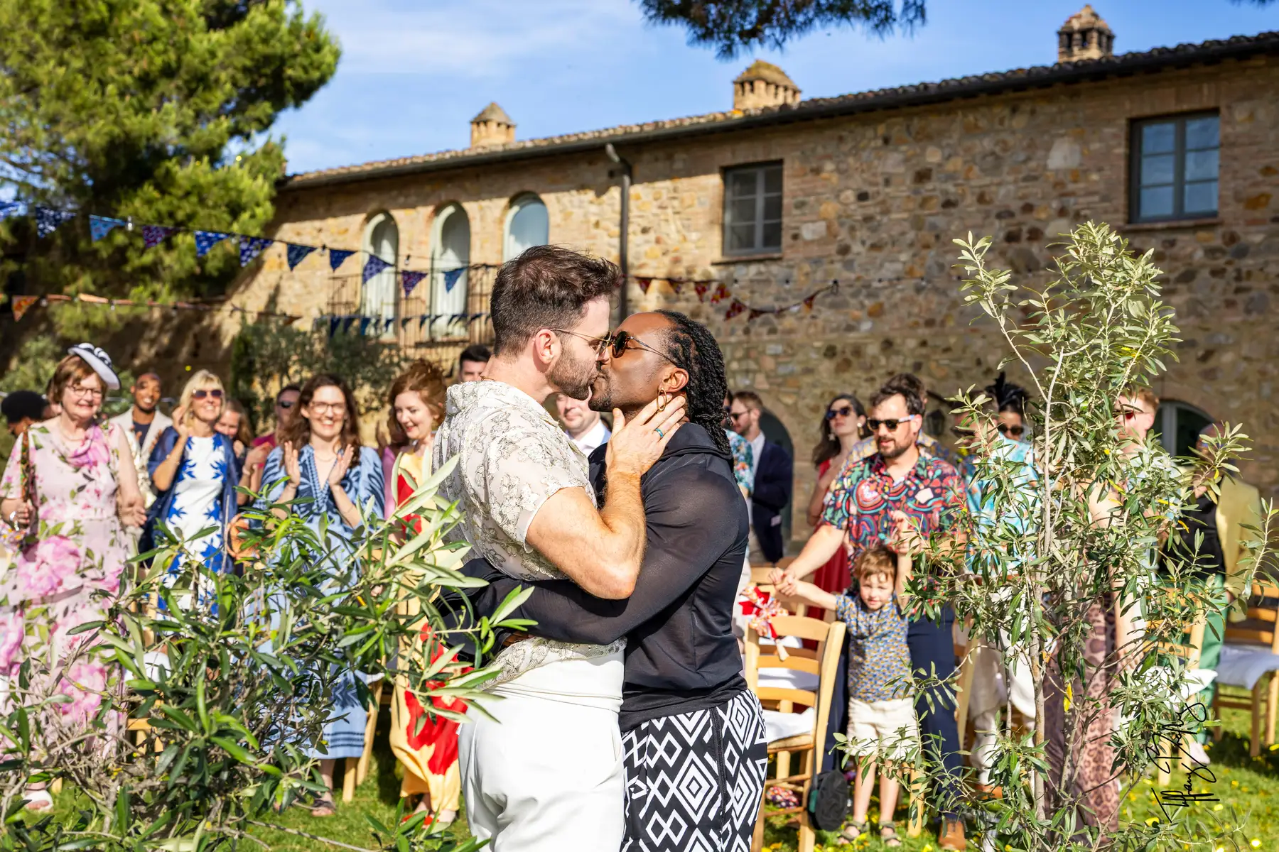 Two grooms share their first kiss during an outdoor ceremony at Conti di San Bonifacio Wine Resort in Tuscany as guests cheer.