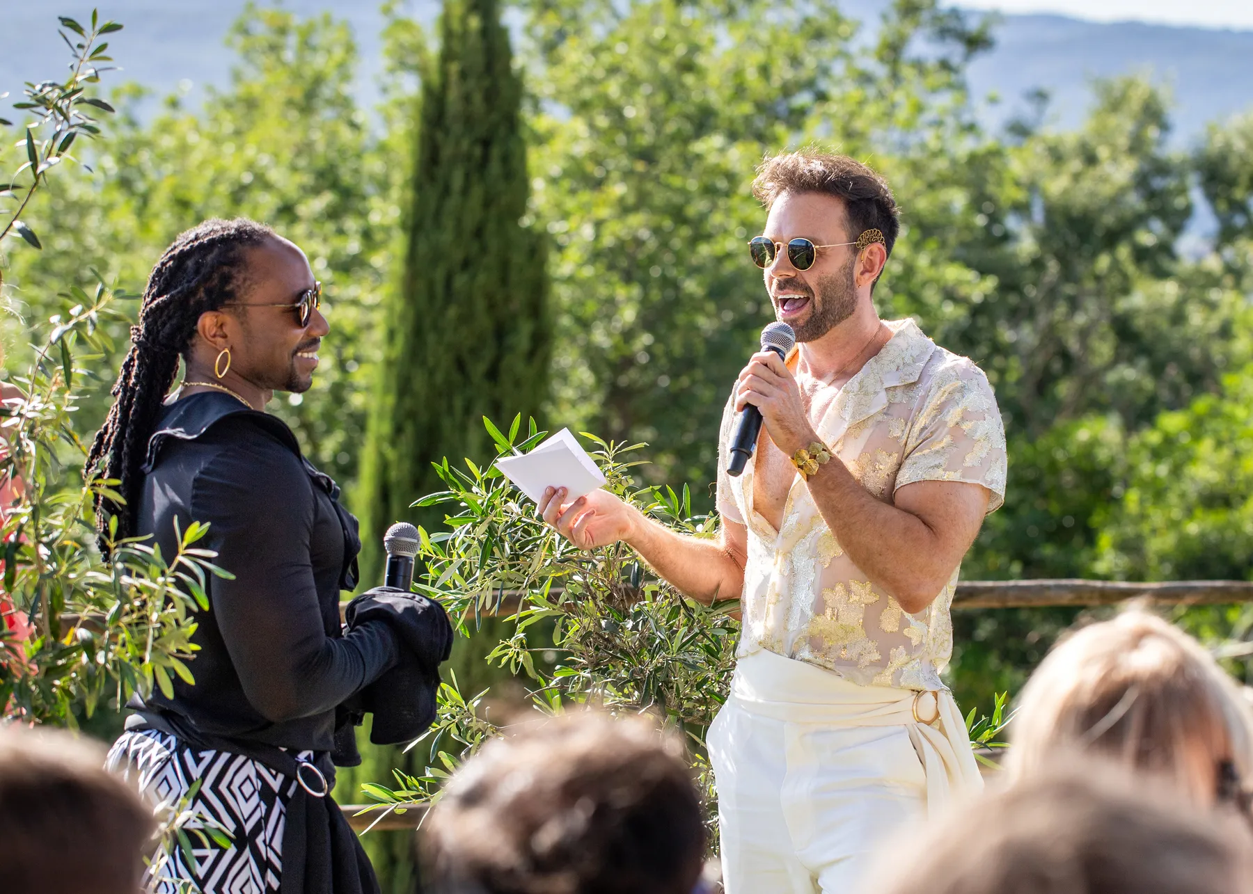 Two grooms exchange vows outdoors at Conti di San Bonifacio Wine Resort in Tuscany, one in white and gold, one in black.