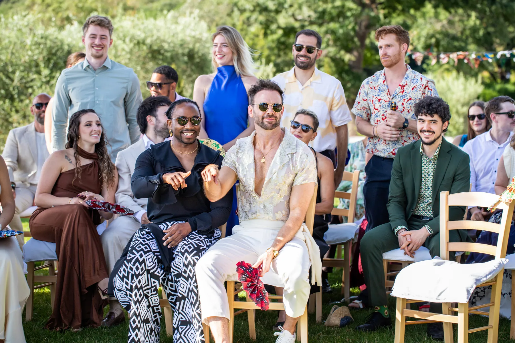 Smiling wedding guests seated outdoors at Conti di San Bonifacio Wine Resort in Tuscany cheer during a same-sex wedding ceremony.