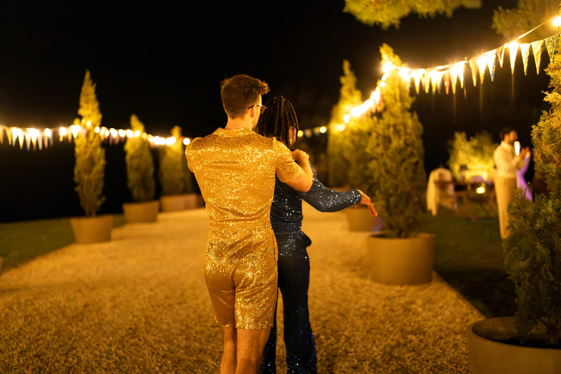 Two grooms in gold and navy sequin outfits dance outdoors at night at Conti di San Bonifacio Wine Resort in Tuscany.