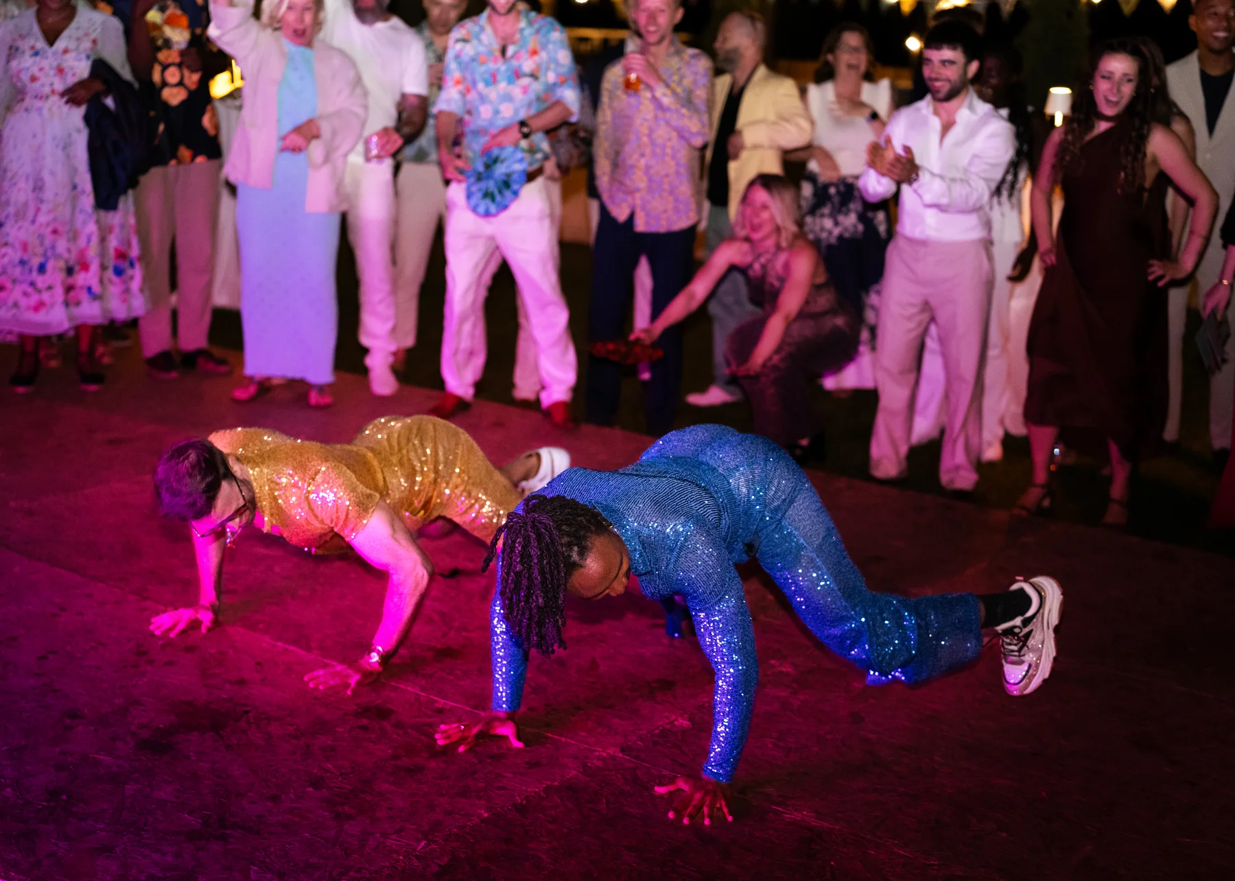 Two grooms in sequined outfits do push-ups on the dance floor as wedding guests cheer at Conti di San Bonifacio in Tuscany.